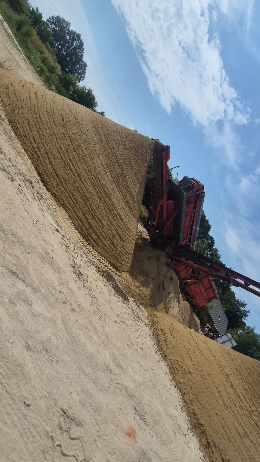 Czerwona przesiewaczka do piasku przy hałdzie piasku na tle błękitnego nieba z chmurami. Widoczne ślady opon na piasku. Obrót zdjęcia o 90 stopni.