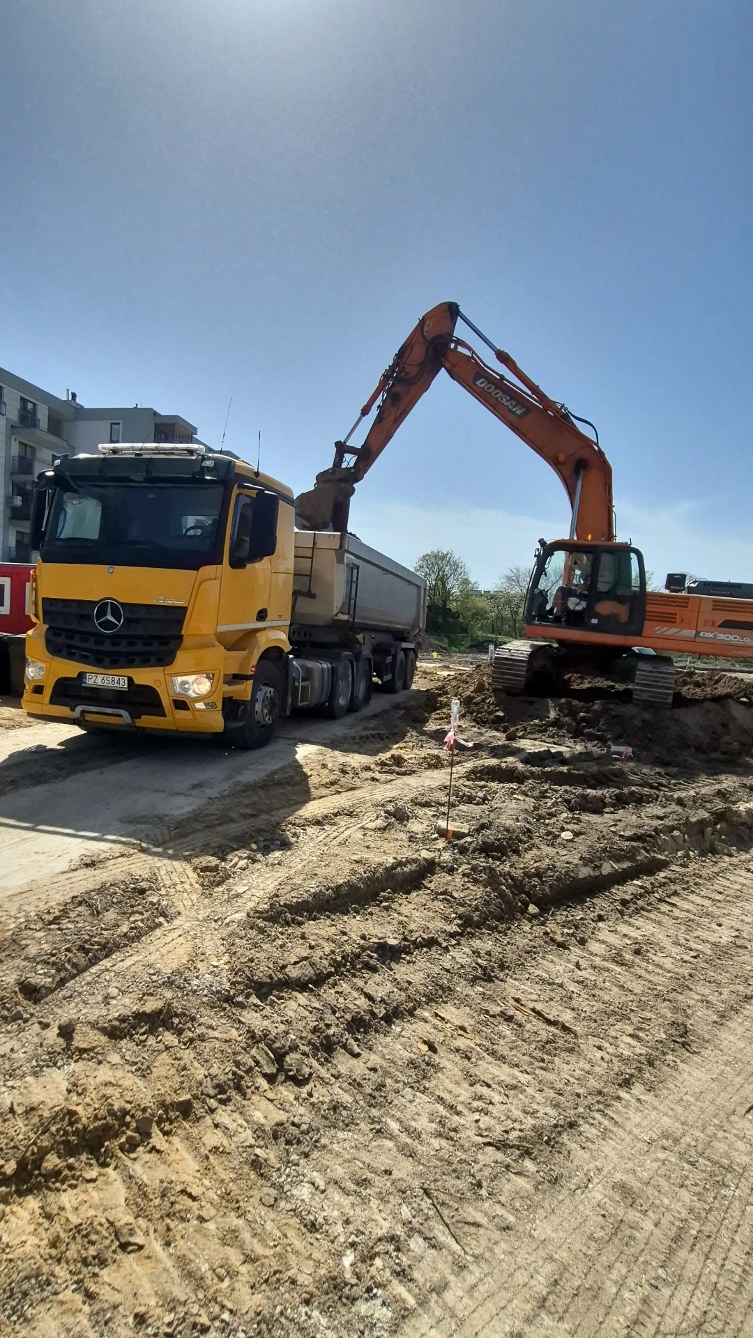 Pomarańczowa koparka Doosan ładuje ziemię do żółtej ciężarówki Mercedes-Benz na placu budowy w słoneczny dzień. Widoczne ślady opon na ziemi i budynek w tle.