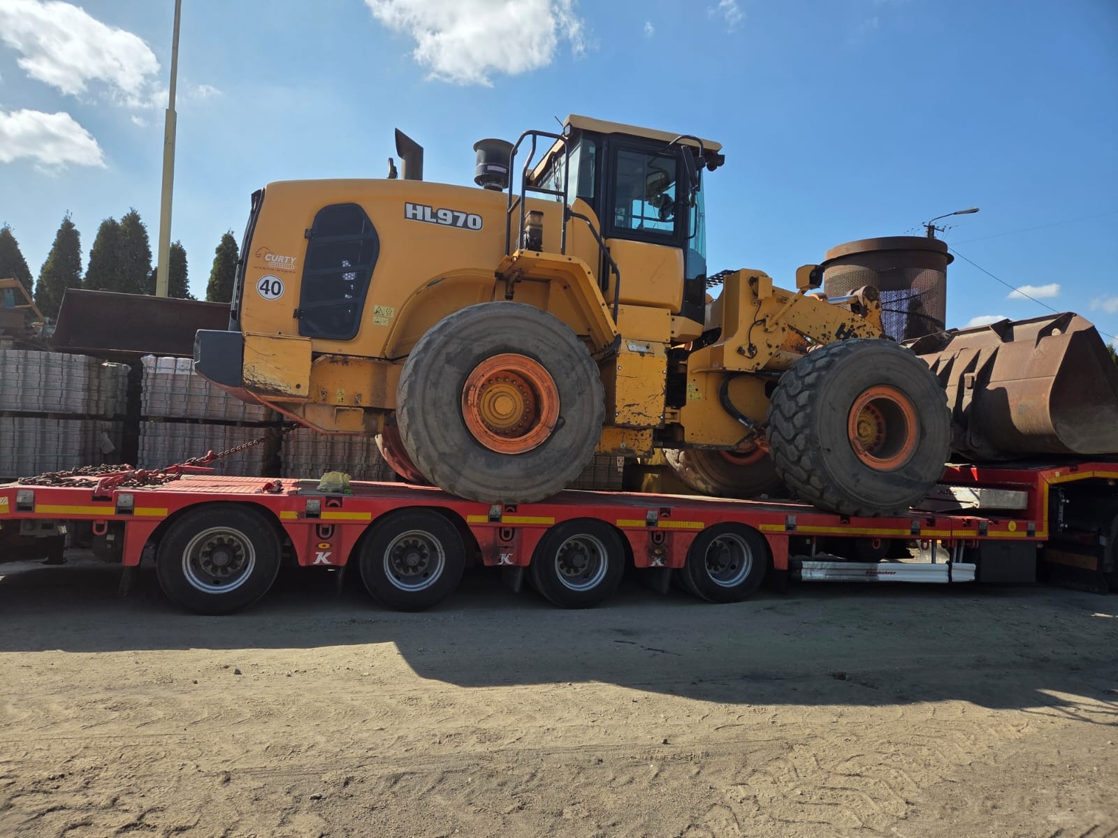 Żółta ładowarka kołowa HL970 na czerwonej lawecie transportowej. Widoczne duże koła, kabina operatora i fragment otoczenia. Transport ciężkiego sprzętu budowlanego.
