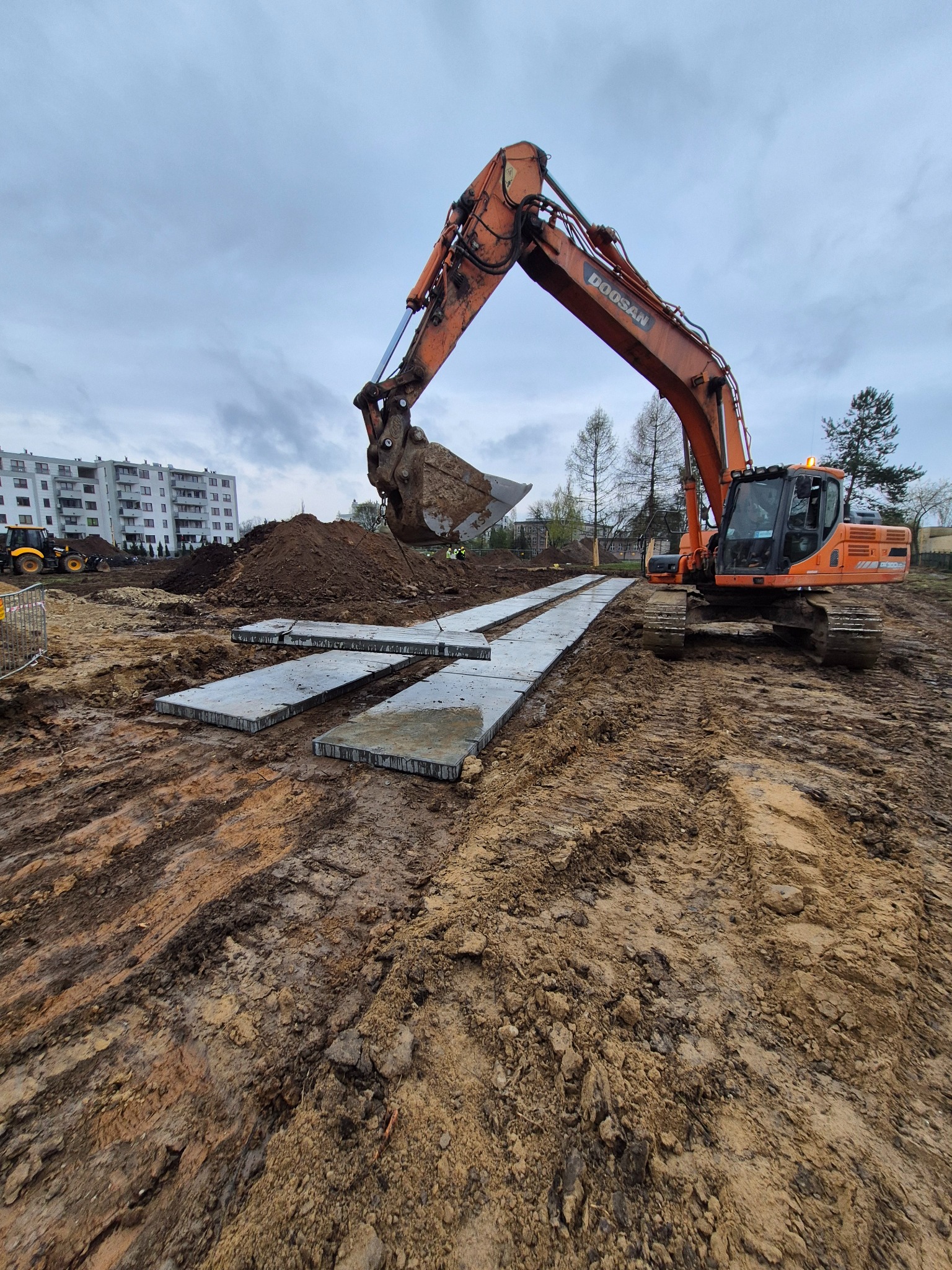 Pomarańczowa koparka Doosan na placu budowy, z łyżką pełną ziemi, w tle budynek mieszkalny i ułożone płyty betonowe na błotnistym terenie.