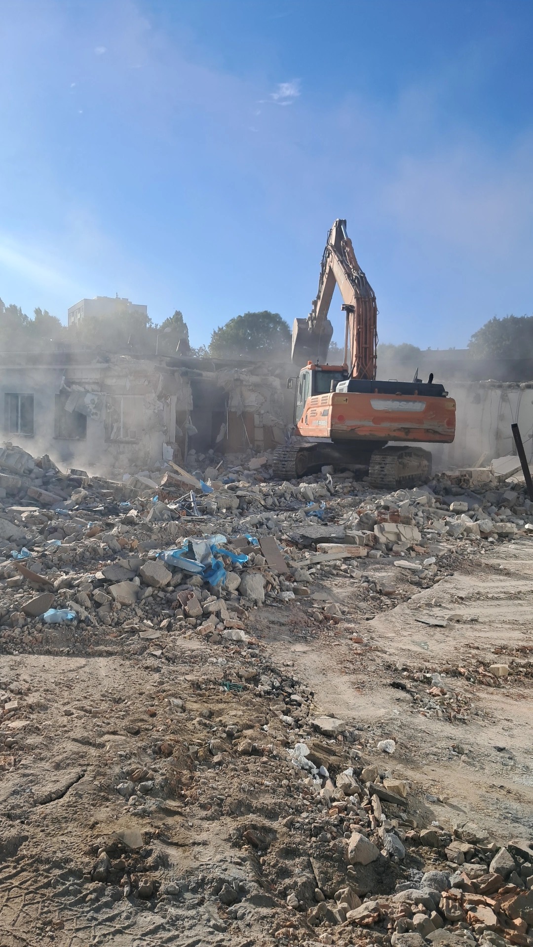 Pomarańczowa koparka na gruzowisku podczas rozbiórki budynku, w tle budynki i drzewa, w powietrzu unosi się pył, widok z dołu.