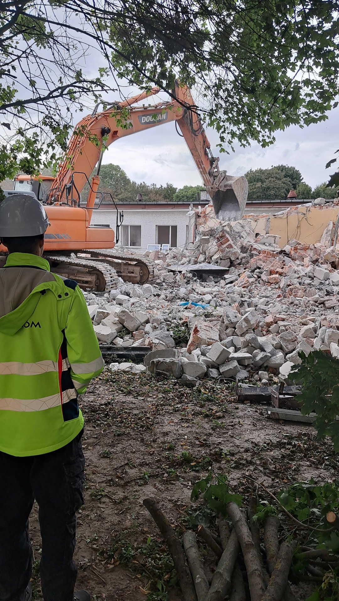 Pomarańczowa koparka Doosan na gruzowisku po rozbiórce budynku. Operator w kasku i kamizelce obserwuje prace. Fragmenty drzew w tle.