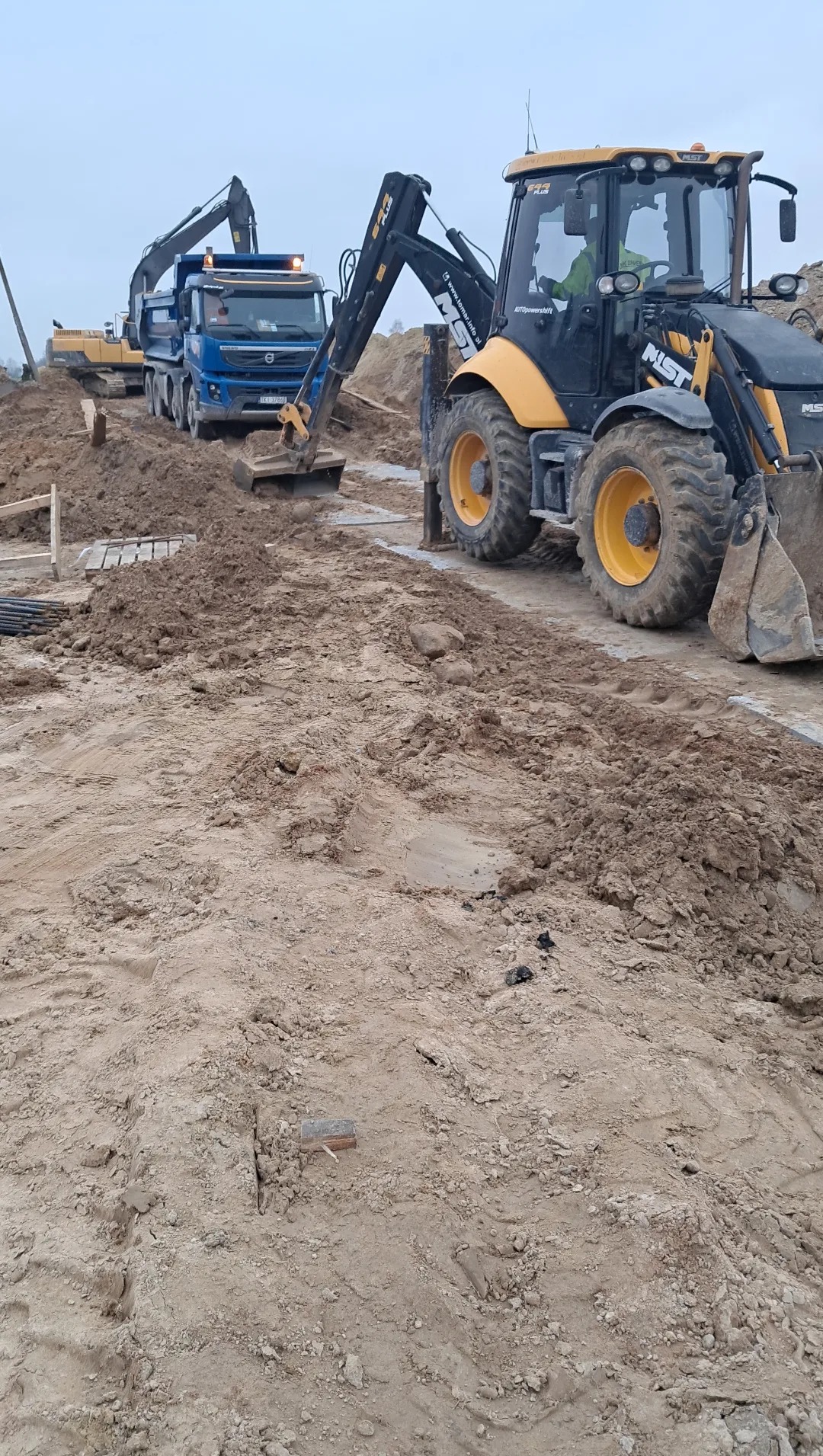 Prace ziemne: koparka JCB, wywrotka Volvo i koparka gąsienicowa na placu budowy. W tle widoczne przygotowane elementy konstrukcyjne.