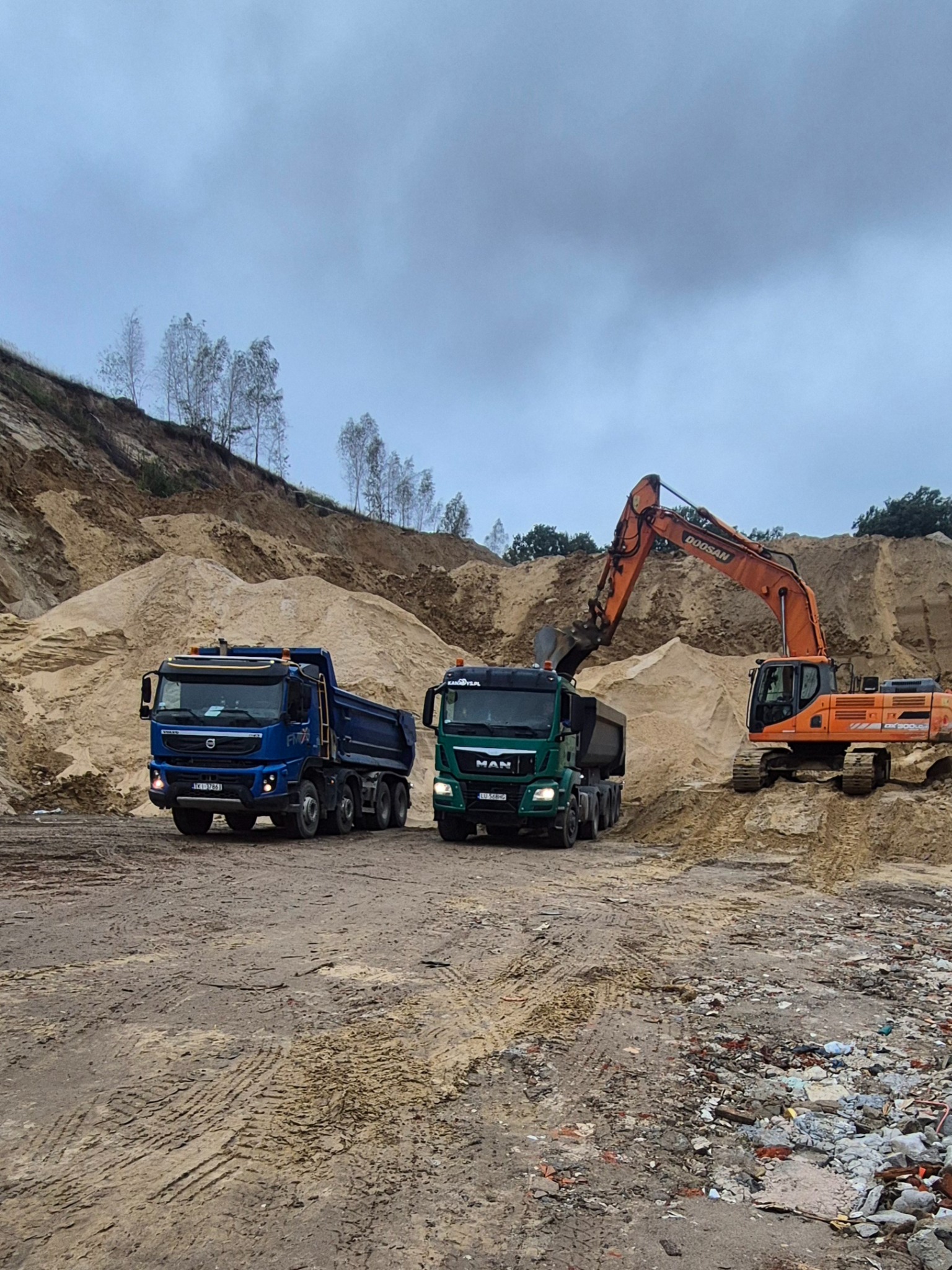 Pomarańczowa koparka Doosan ładuje urobek do zielonej ciężarówki MAN, obok stoi niebieska ciężarówka Volvo, w tle hałda piasku i pochmurne niebo.