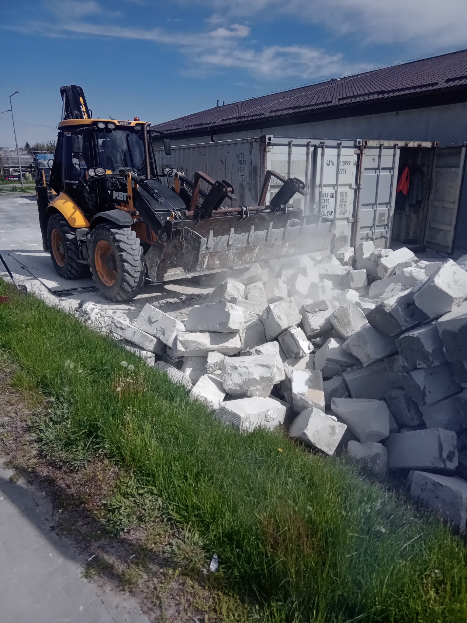 Żółto-czarna koparko-ładowarka MST nabiera gruz z rozbiórki obok kontenera. Praca w toku, widoczna trawa i fragment nieba.
