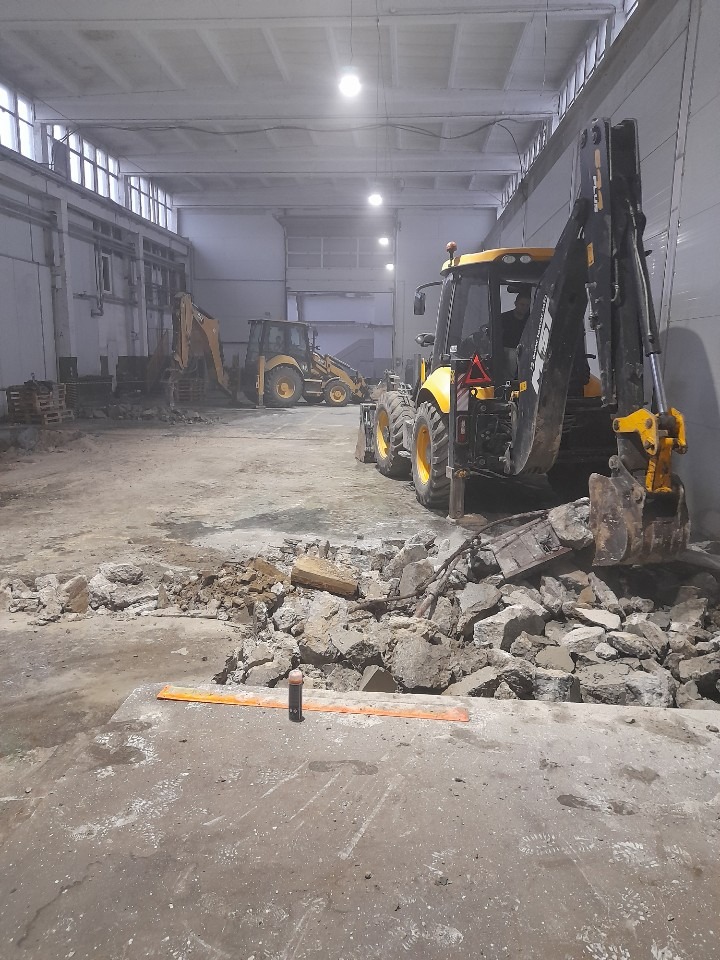 Wnętrze hali w trakcie rozbiórki z dwoma żółtymi koparkami. Gruz na podłodze, pomarańczowa listwa i butelka w tle. Widoczne ślady opon i stóp.