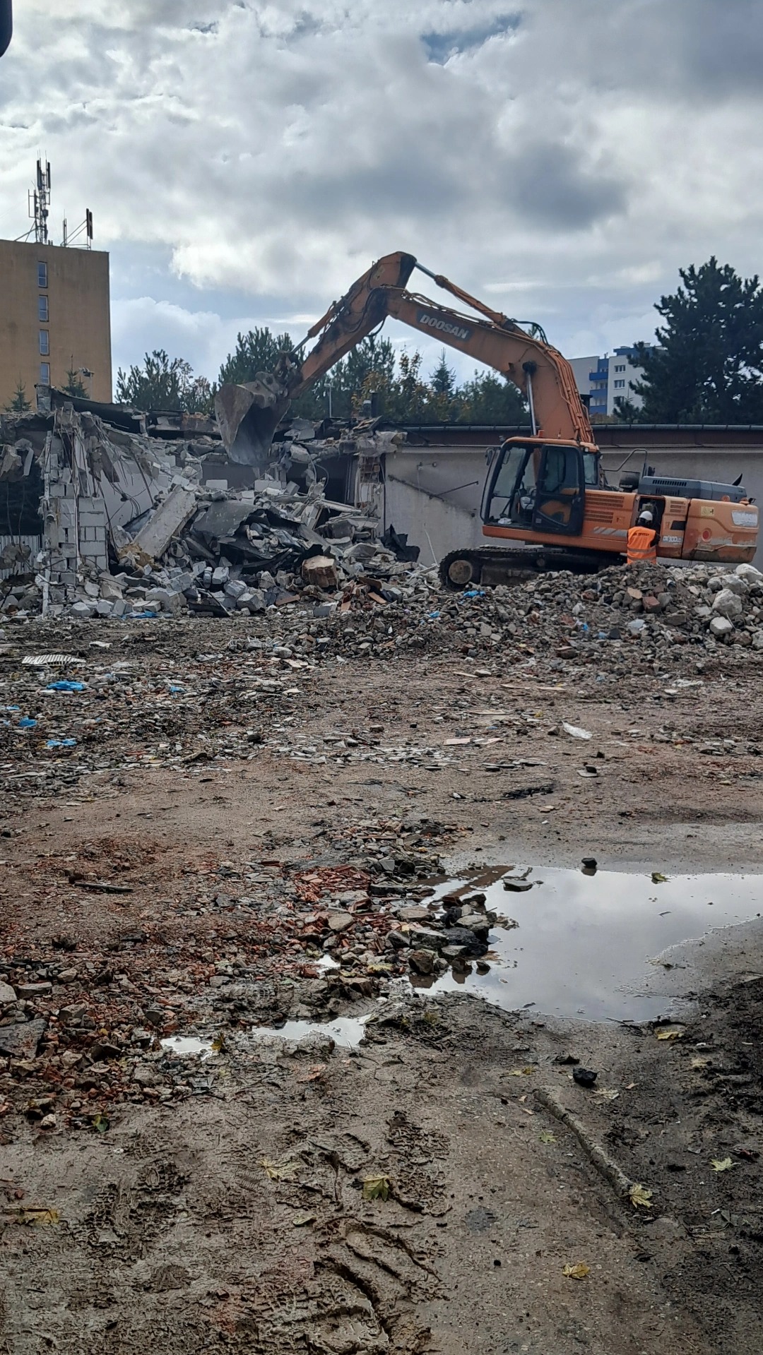 Pomarańczowa koparka Doosan podczas rozbiórki budynku. Na pierwszym planie gruz i błoto. W tle budynki i pochmurne niebo. Widoczny operator w kamizelce.