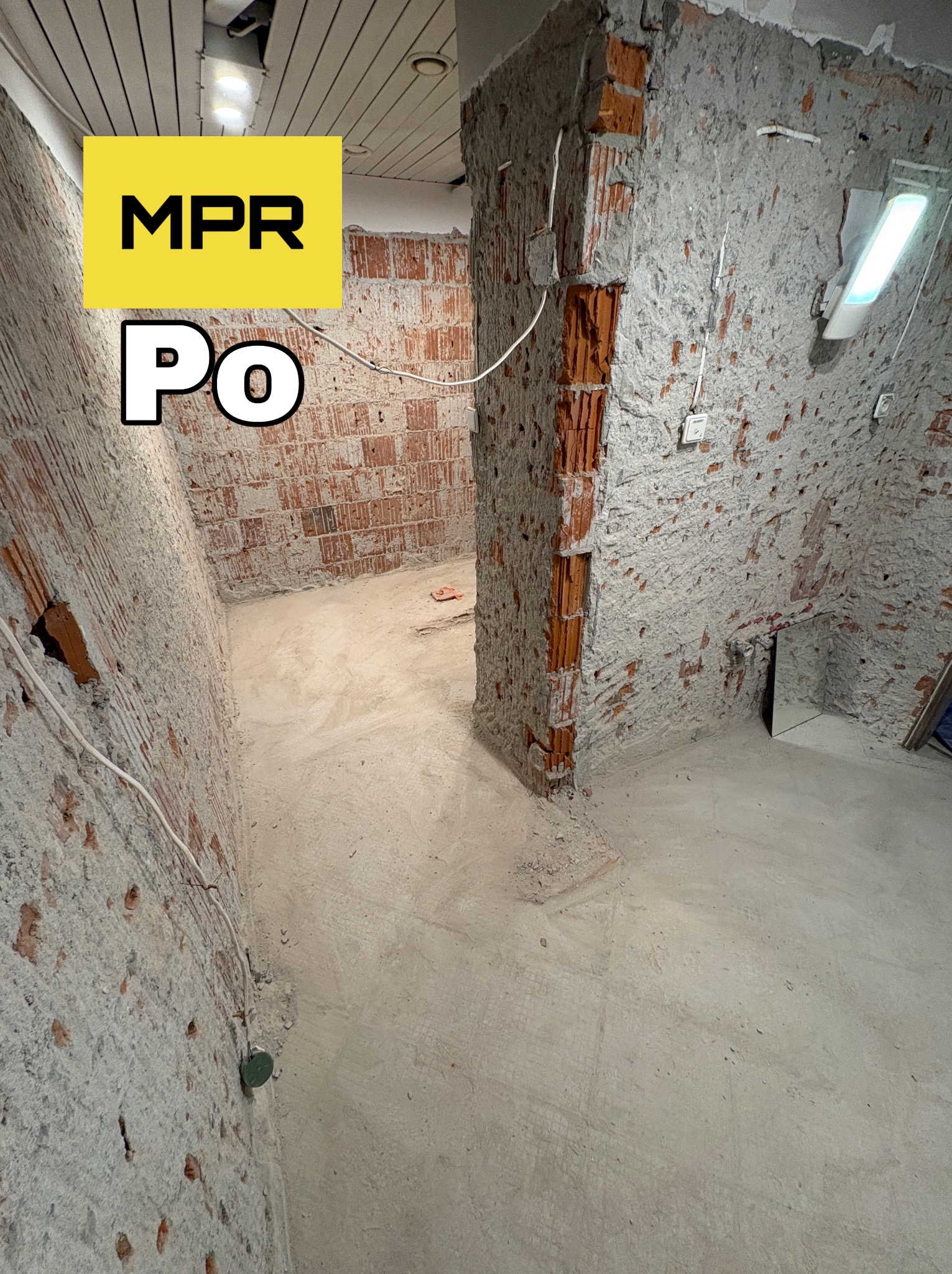 Wnętrze po skuciu tynków i usunięciu części ściany, widoczne odsłonięte cegły i instalacje. Na ścianie logo firmy MPR i napis 'Po'.
