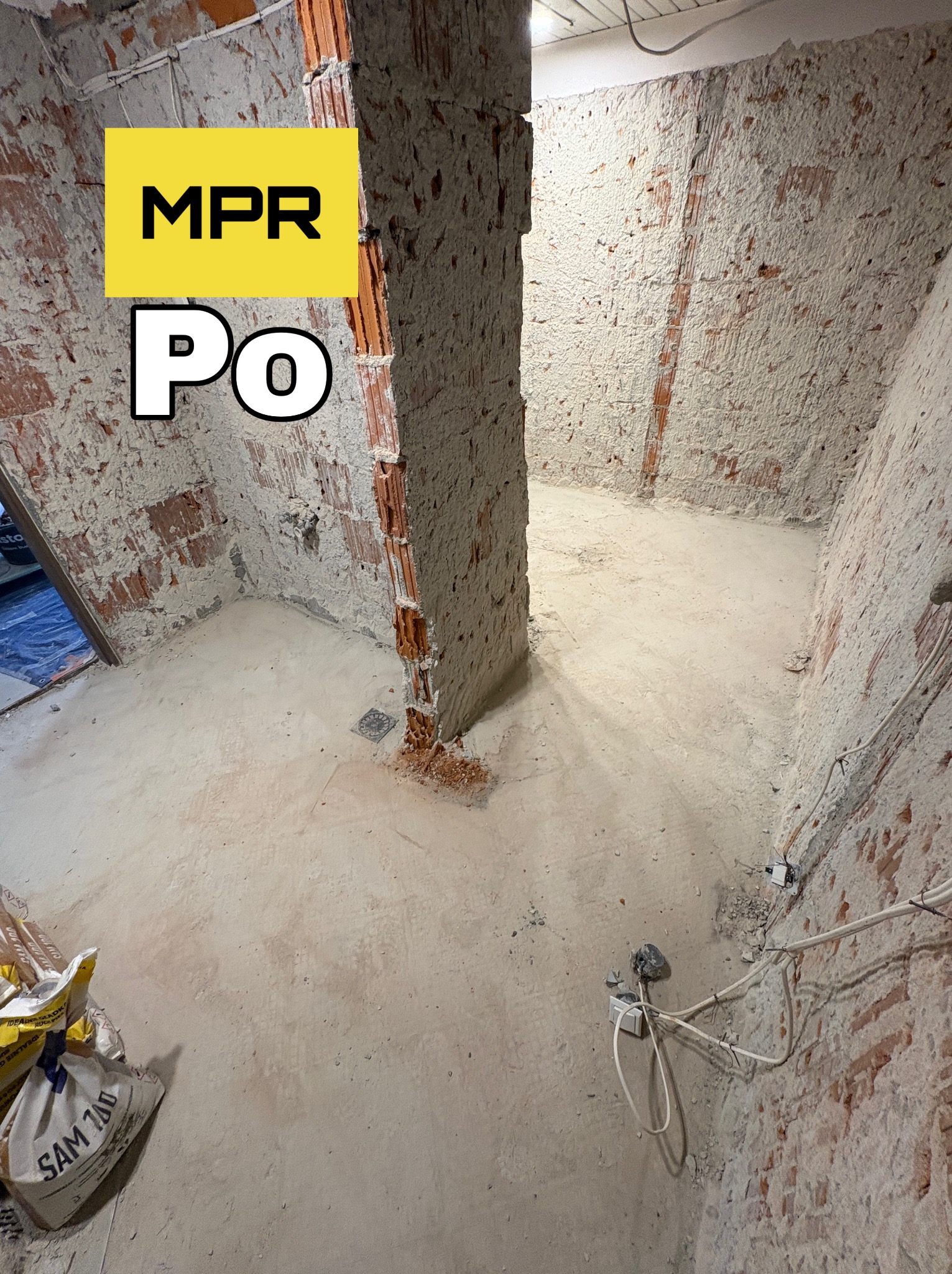 Wnętrze po usunięciu ściany działowej, widoczne odsłonięte cegły i resztki zaprawy. Na podłodze worki z zaprawą. Odsłonięte kable elektryczne.