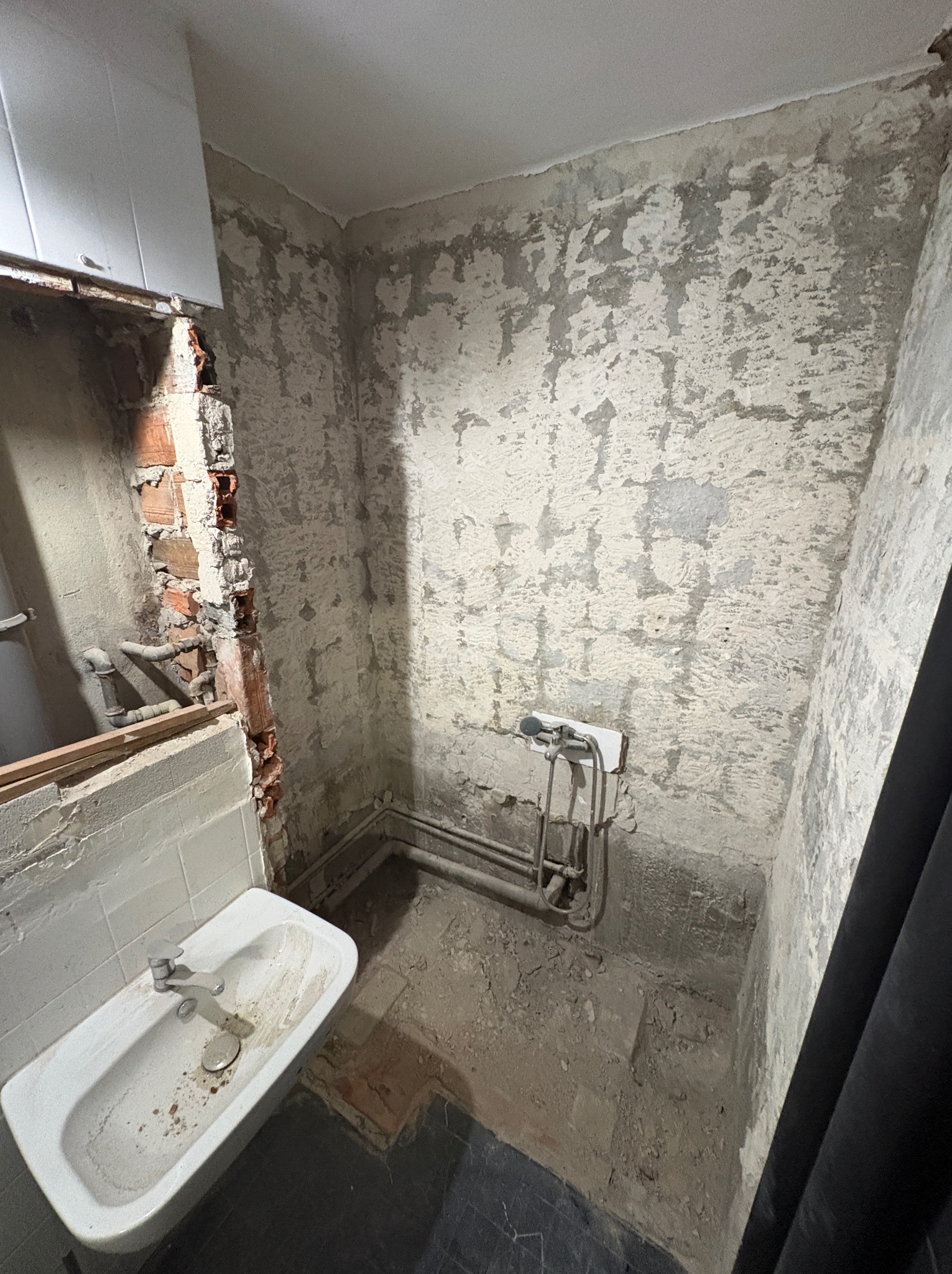 Łazienka w trakcie demontażu: odsłonięta cegła po wyburzeniu, stara umywalka, rury i ściany z resztkami farby. Widok z góry, surowy stan przed remontem.