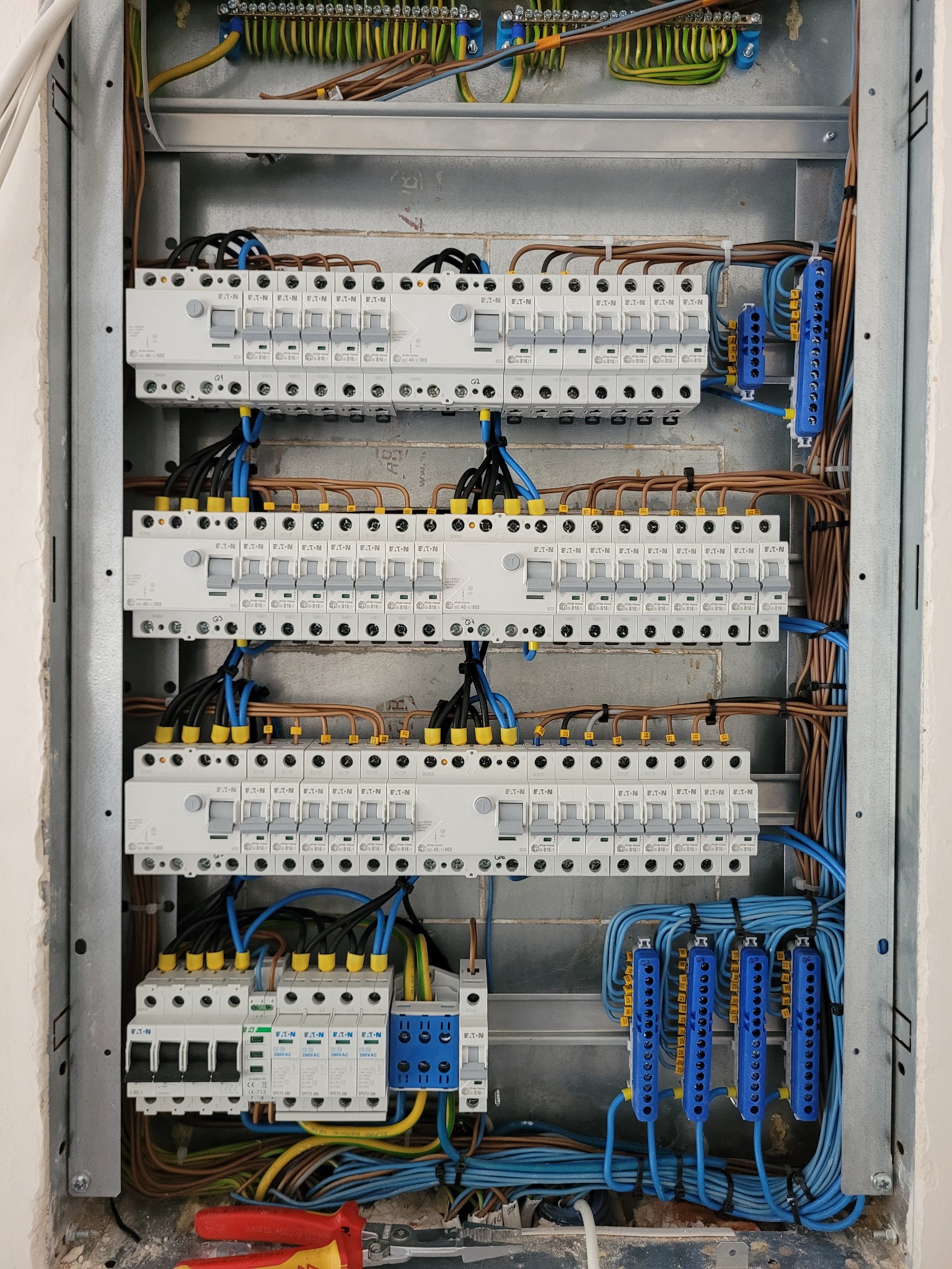 Rozdzielnia elektryczna w metalowej obudowie z widocznymi bezpiecznikami, przewodami w kolorach brązowym i niebieskim oraz narzędziami na dole.