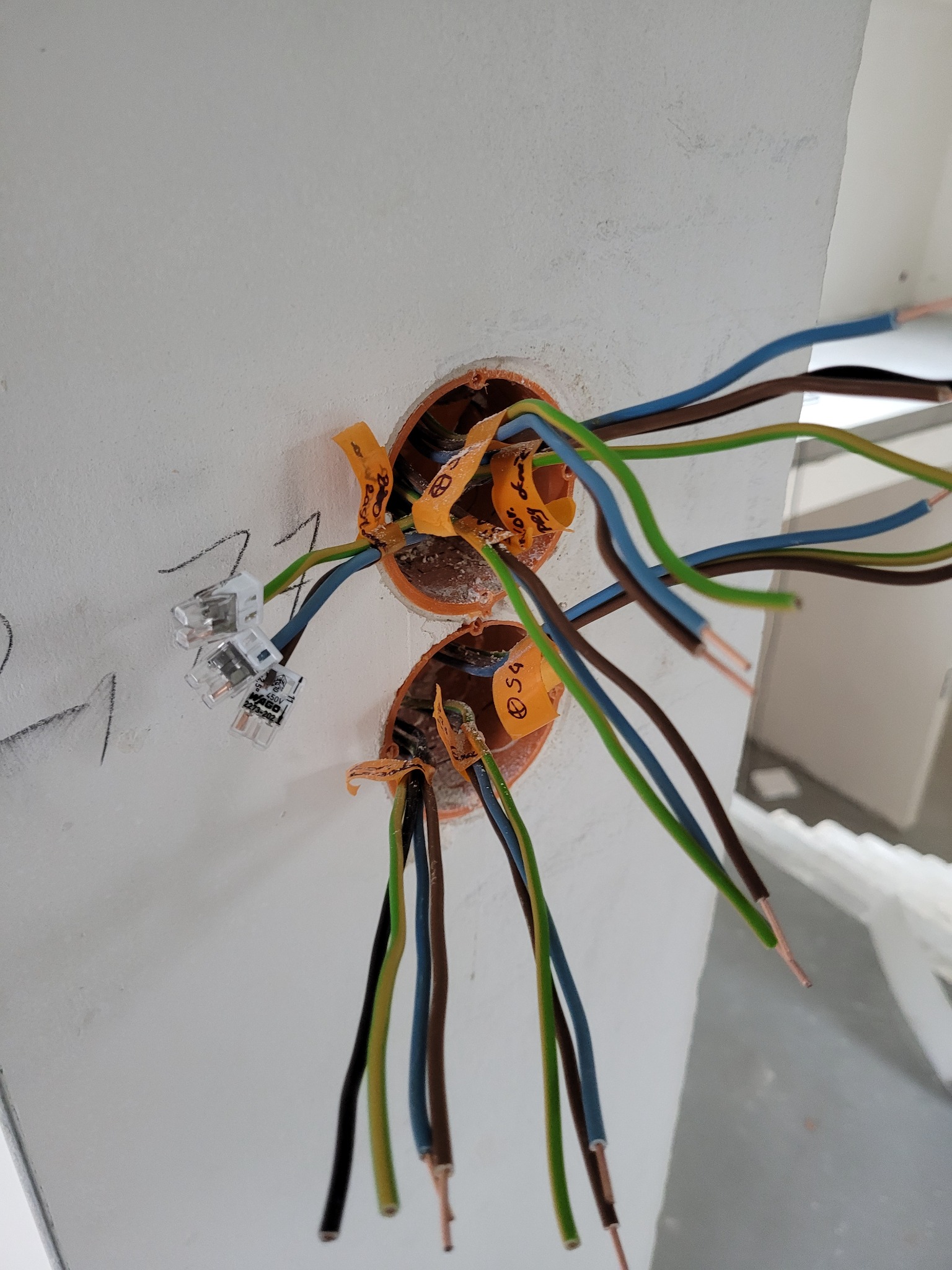 Dwa otwory w ścianie z wystającymi przewodami elektrycznymi o różnych kolorach, zabezpieczonymi pomarańczową taśmą i złączkami WAGO. Widoczne oznaczenia na taśmach.