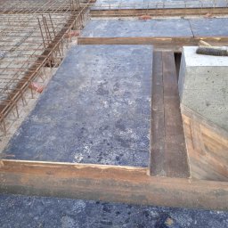 RAFTECH - Szczegółowe ujęcie szalunku stropowego z widocznym zbrojeniem i betonowym słupem, przygotowanego do wylania betonu. Widoczne deski i metalowe elementy konstrukcyjne.