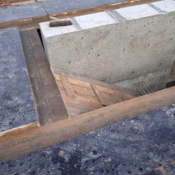 RAFTECH - Szczegółowy widok szalunku fundamentowego z betonu i drewnianych elementów konstrukcyjnych, ukazujący etap przygotowania do wylewania betonu. Widoczna szczotka na betonowym elemencie.