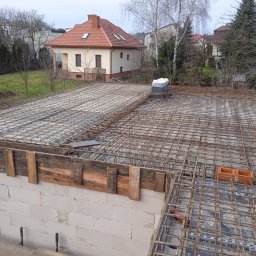 RAFTECH - Zbrojenie stropu domu jednorodzinnego z bloczków, widoczne szalunki i siatka zbrojeniowa. W tle dom z czerwonym dachem i drzewa bez liści.