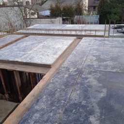 RAFTECH - Szare płyty szalunkowe i zbrojenie na budowie domu, widok z góry. W tle zabudowa miejska i drzewa bez liści. Etap przygotowania do wylewania stropu.