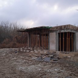 RAFTECH - Surowy stan domu z betonu komórkowego w trakcie budowy. Widoczne podpory stropu i szalunki. Teren wokół placu budowy jest błotnisty, z porozrzucanymi elementami budowlanymi.