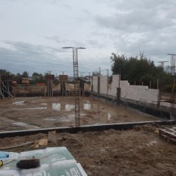 RAFTECH - Fundamenty domu w Tomaszowie Lubelskim w trakcie budowy. Widoczne zbrojenia, częściowo wymurowane ściany fundamentowe i sprzęt budowlany w tle. Pochmurny dzień.