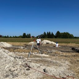 RAFTECH - Prace geodezyjne przy wykopie pod fundamenty domu jednorodzinnego w słoneczny dzień. Widoczny niwelator na statywie i pracownicy w trakcie pomiarów.