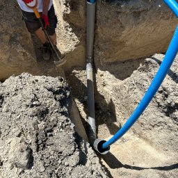 RAFTECH - Wykop z ułożonymi rurami kanalizacyjnymi, widoczny fragment niebieskiego węża i pracownik z łopatą. Ujęcie z góry, ostre światło słoneczne.