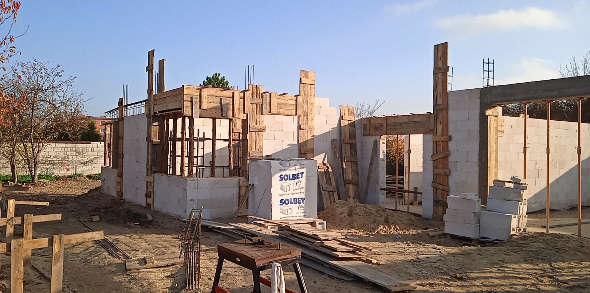 Budowa domu jednorodzinnego w trakcie wznoszenia ścian z bloczków SOLBET. Widoczne drewniane szalunki i zbrojenia. Słoneczny dzień, błękitne niebo w tle.