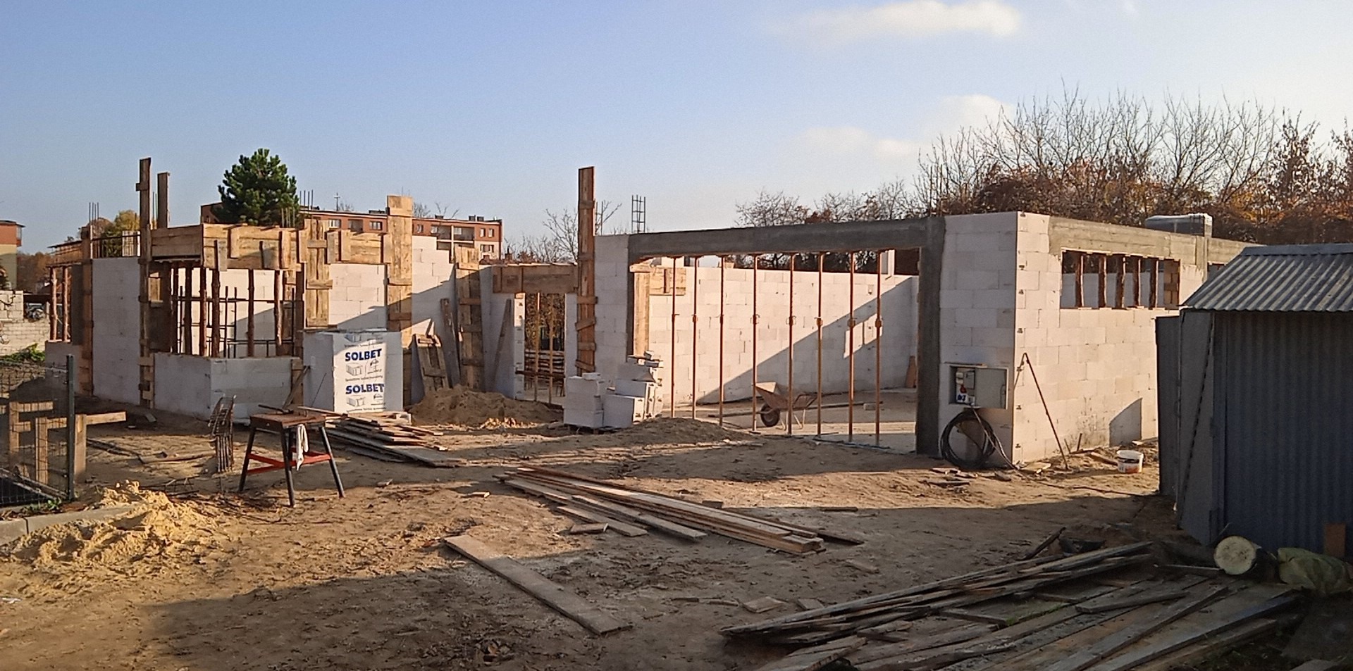 Budowa domu jednorodzinnego w trakcie realizacji: wzniesione ściany z betonu komórkowego Solbet, widoczne szalunki i zbrojenia. Teren budowy z piaskiem i deskami.