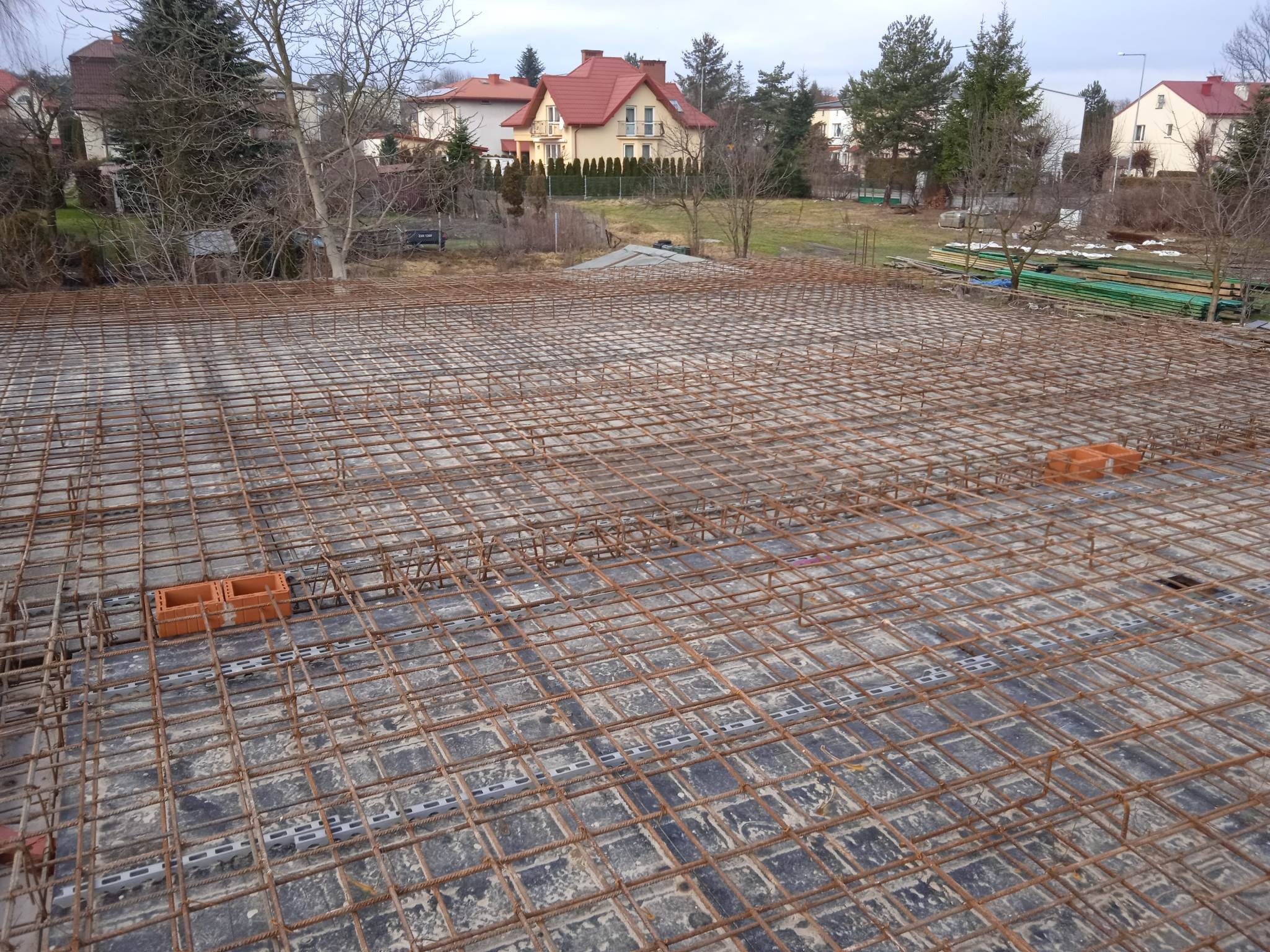 Siatka zbrojeniowa na fundamencie płyty, widok z góry. W tle zabudowa jednorodzinna i drzewa. Elementy dystansowe z tworzywa i cegły.