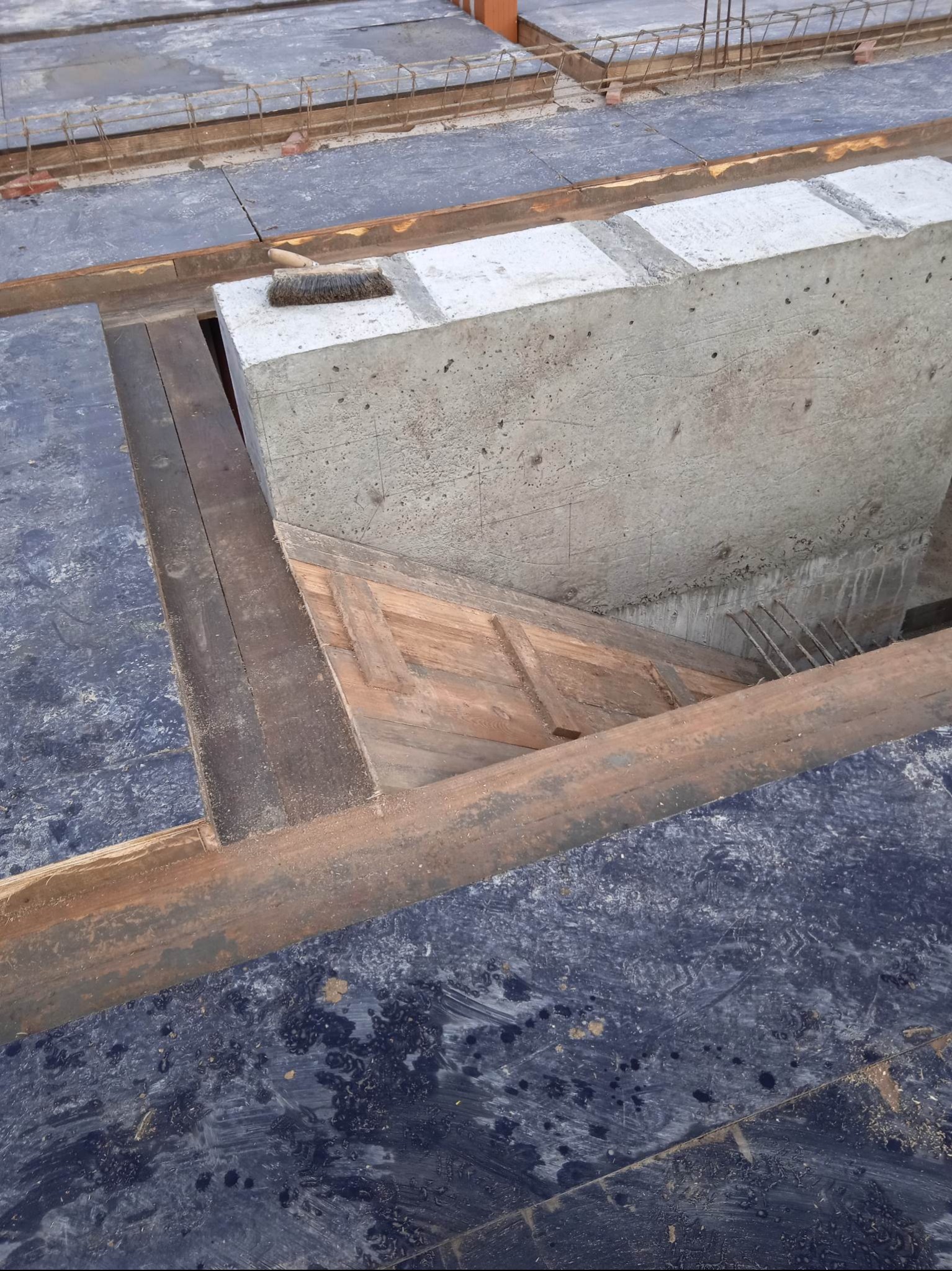 Szczegółowy widok szalunku fundamentowego z betonu i drewnianych elementów konstrukcyjnych, ukazujący etap przygotowania do wylewania betonu. Widoczna szczotka na betonowym elemencie.
