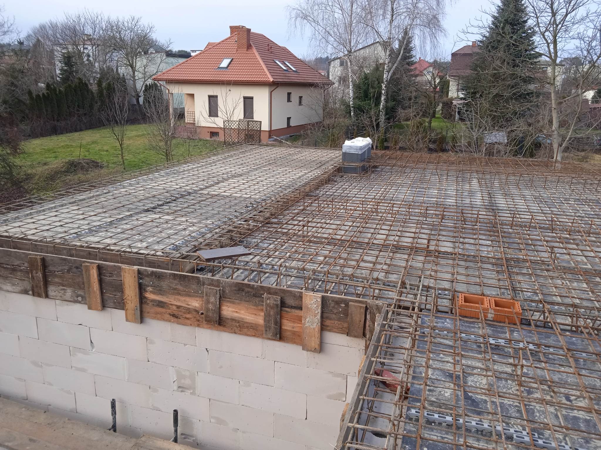 Zbrojenie stropu domu jednorodzinnego z bloczków, widoczne szalunki i siatka zbrojeniowa. W tle dom z czerwonym dachem i drzewa bez liści.