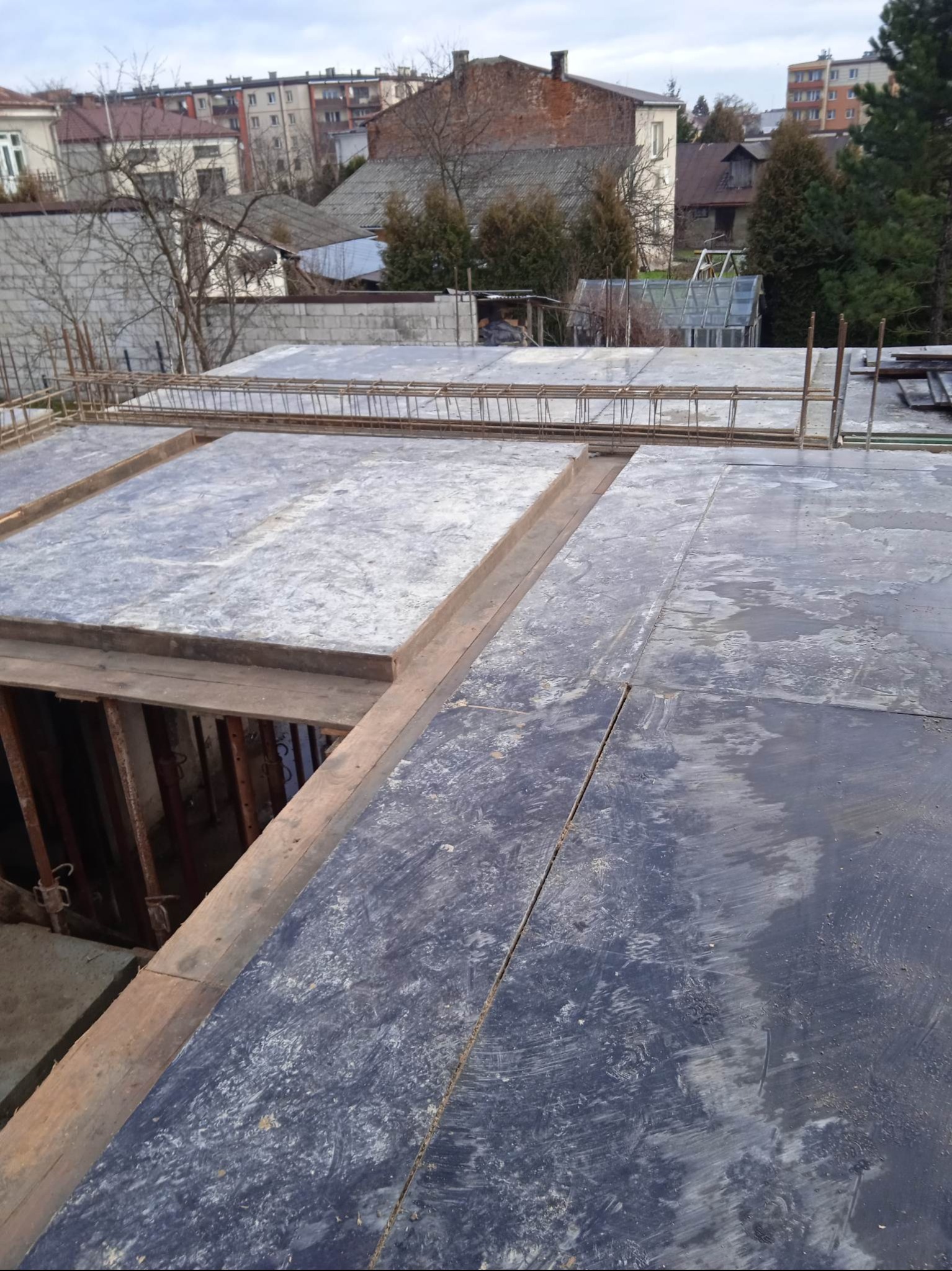 Szare płyty szalunkowe i zbrojenie na budowie domu, widok z góry. W tle zabudowa miejska i drzewa bez liści. Etap przygotowania do wylewania stropu.