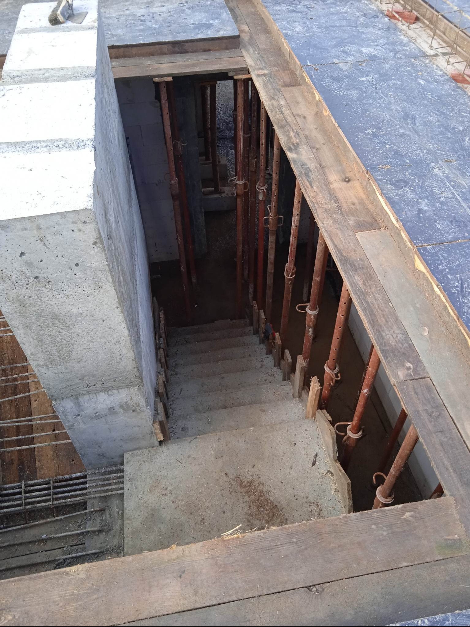 Wykop fundamentów z betonowymi schodami i drewnianym szalunkiem, widok z góry. Stalowe podpory pionowe. Etap budowy domu.