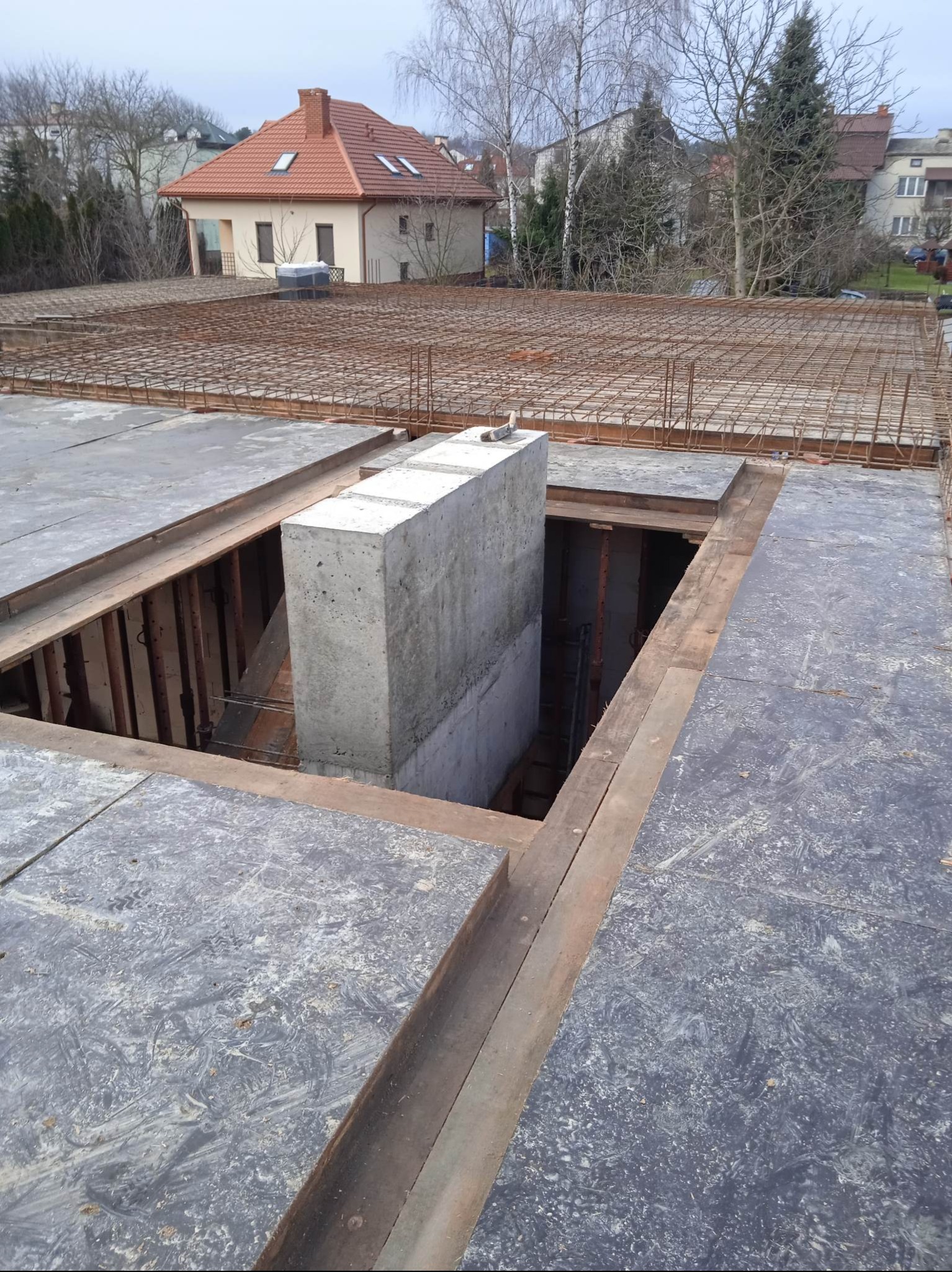 Zbrojenie stropu z otworem na schody i betonowym słupem w surowym stanie. Widoczne deski szalunkowe i siatka z prętów. W tle budynek mieszkalny.