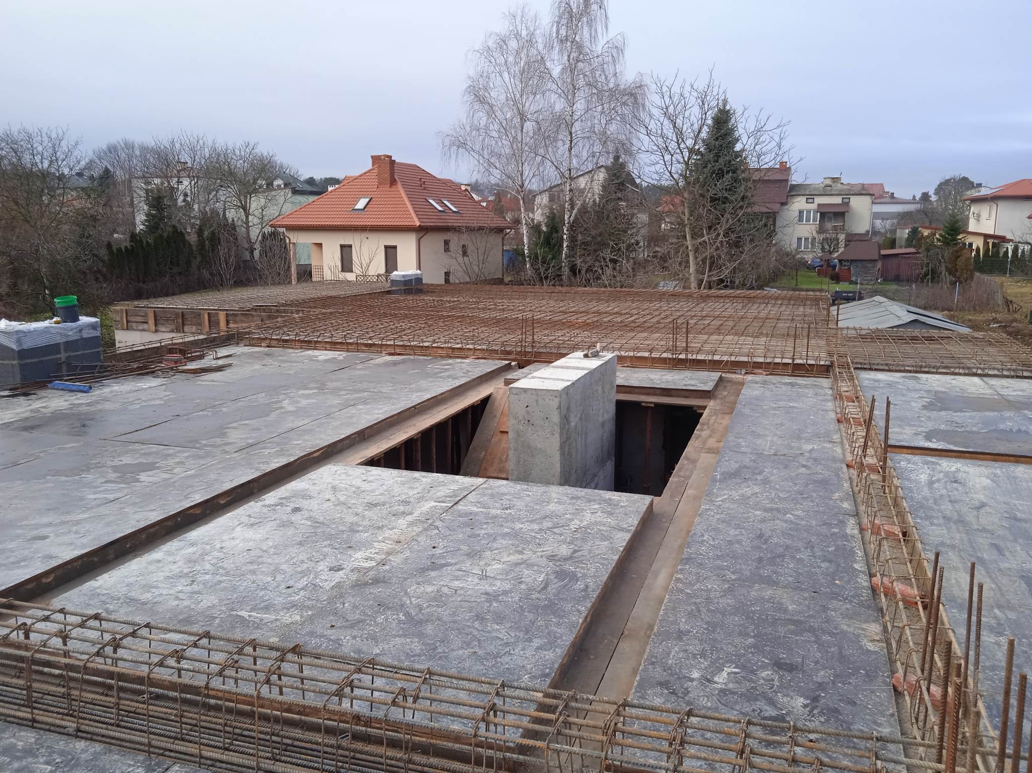 Zbrojenie stropu budynku jednorodzinnego z widocznymi otworami na schody i instalacje, przygotowane do zalania betonem. W tle zabudowa mieszkalna.