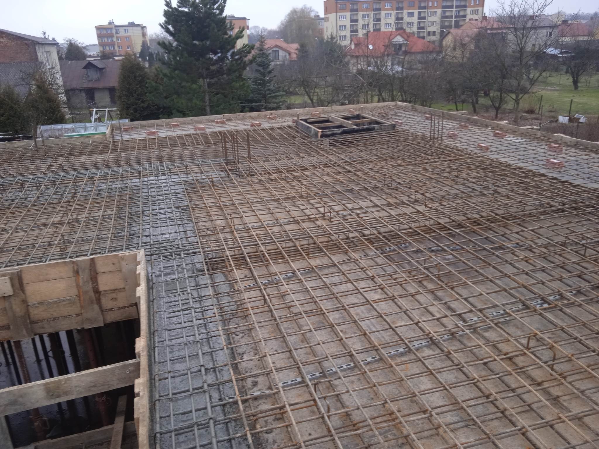Zbrojenie stropu budynku w trakcie budowy, widok z góry. Stalowe pręty tworzą siatkę, częściowo zalane betonem. W tle zabudowa miejska i drzewa.
