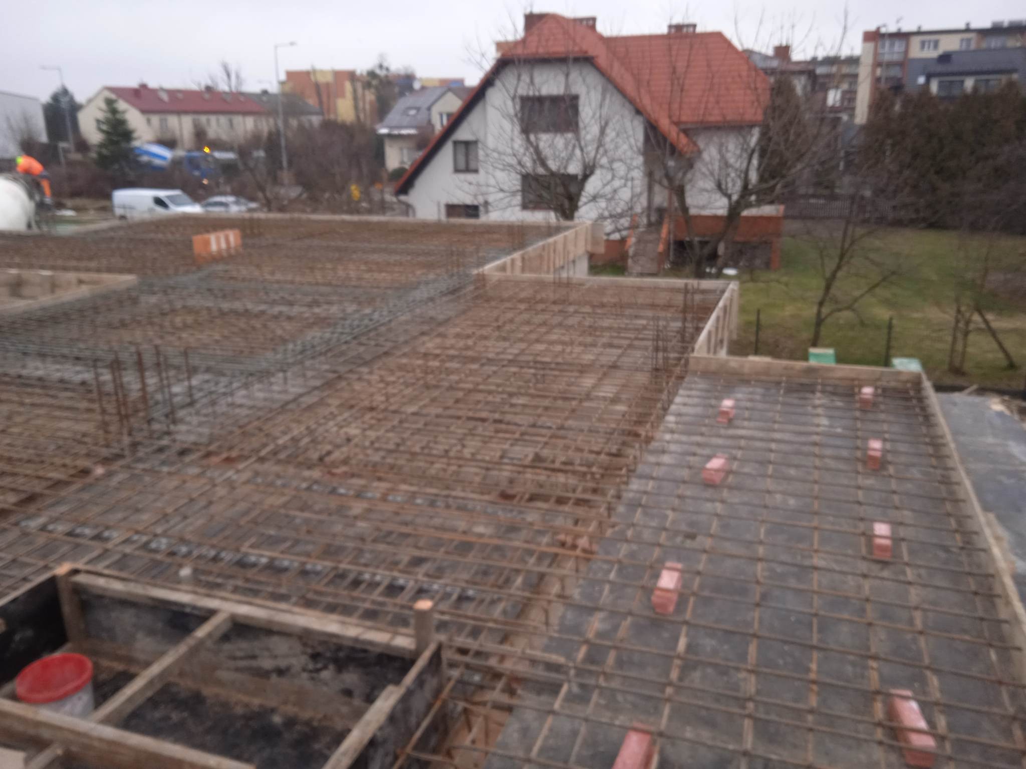 Zbrojenie fundamentów domu jednorodzinnego, widok z góry na siatkę zbrojeniową i szalunki. W tle budynki mieszkalne i sprzęt budowlany.