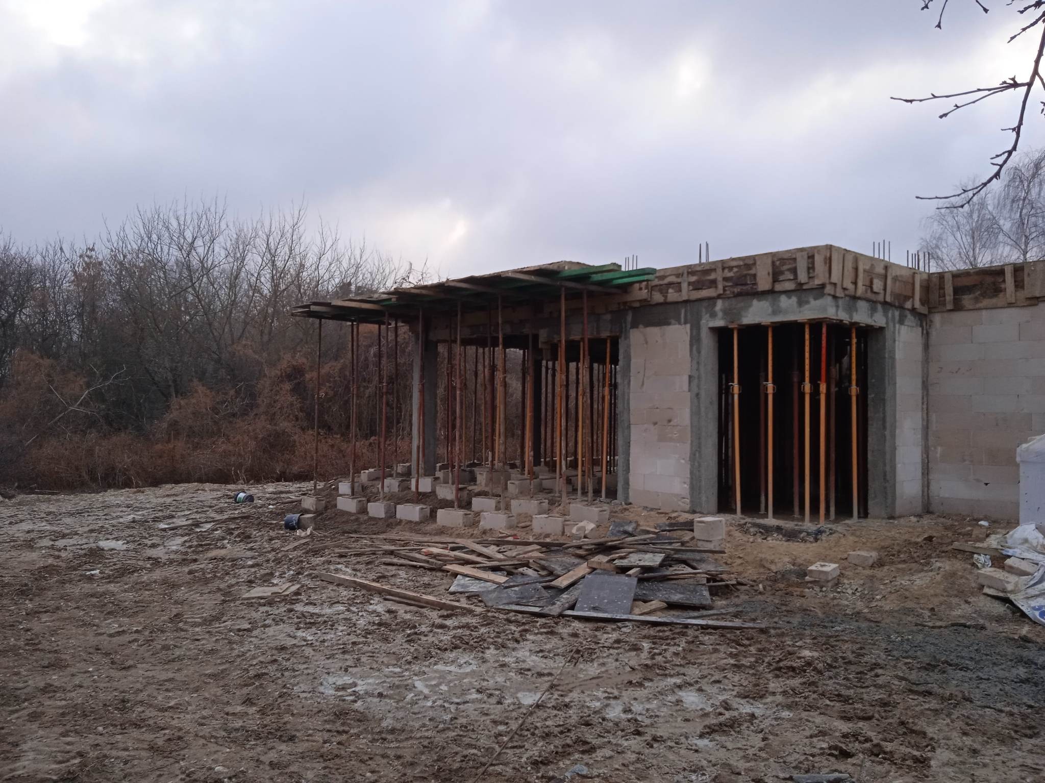 Surowy stan domu z betonu komórkowego w trakcie budowy. Widoczne podpory stropu i szalunki. Teren wokół placu budowy jest błotnisty, z porozrzucanymi elementami budowlanymi.