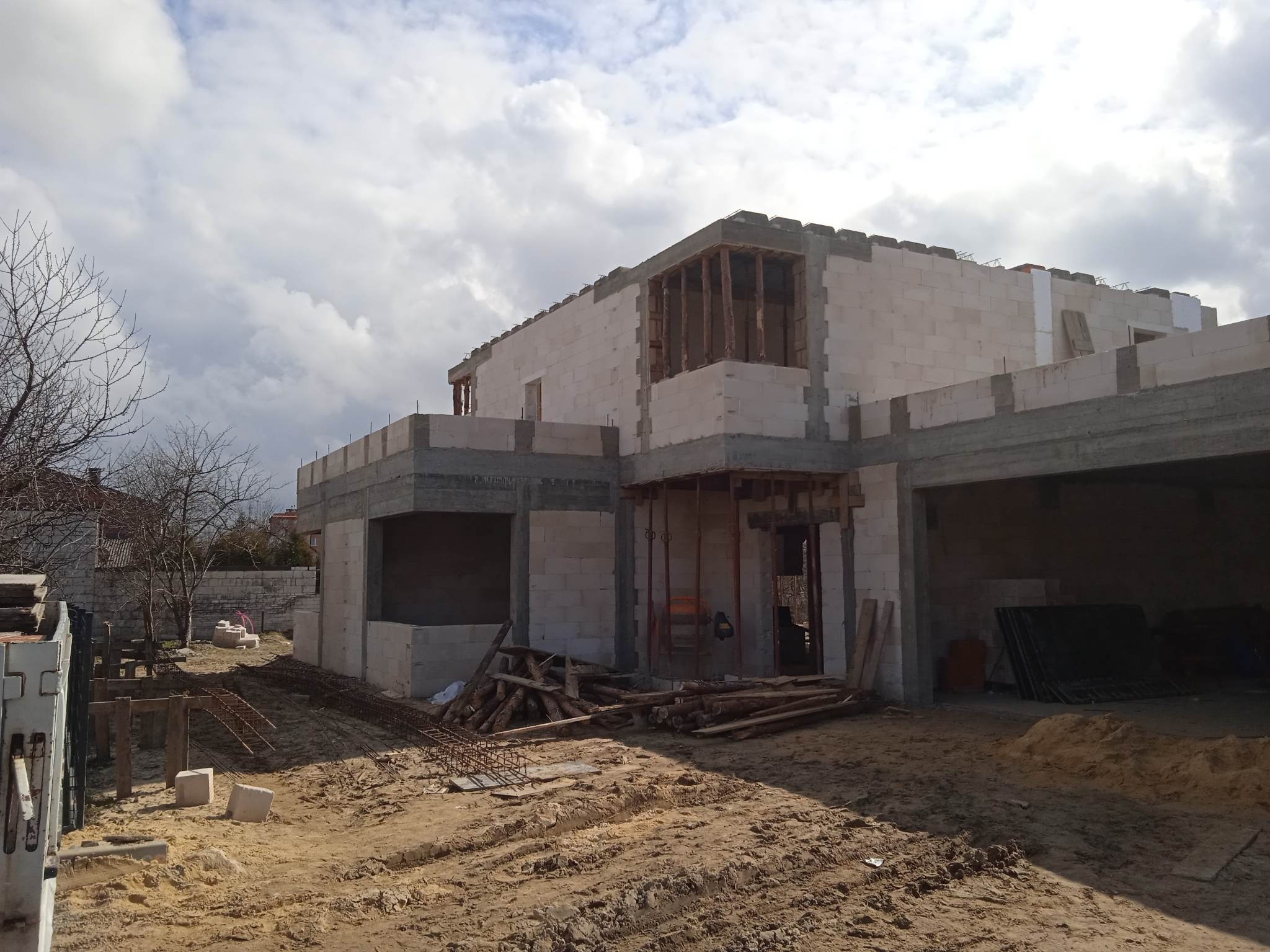 Surowy stan domu z betonu komórkowego w trakcie budowy. Widoczne zbrojenia, drewniane podpory i elementy szalunków. Teren budowy z piaskiem i materiałami budowlanymi.