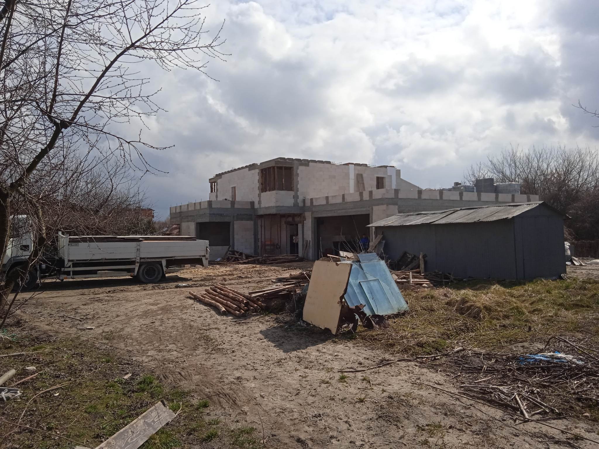 Surowy stan budowy domu z betonu komórkowego, widoczny z zewnątrz, z ciężarówką w tle i stertą drewna na pierwszym planie. Niebo pochmurne.