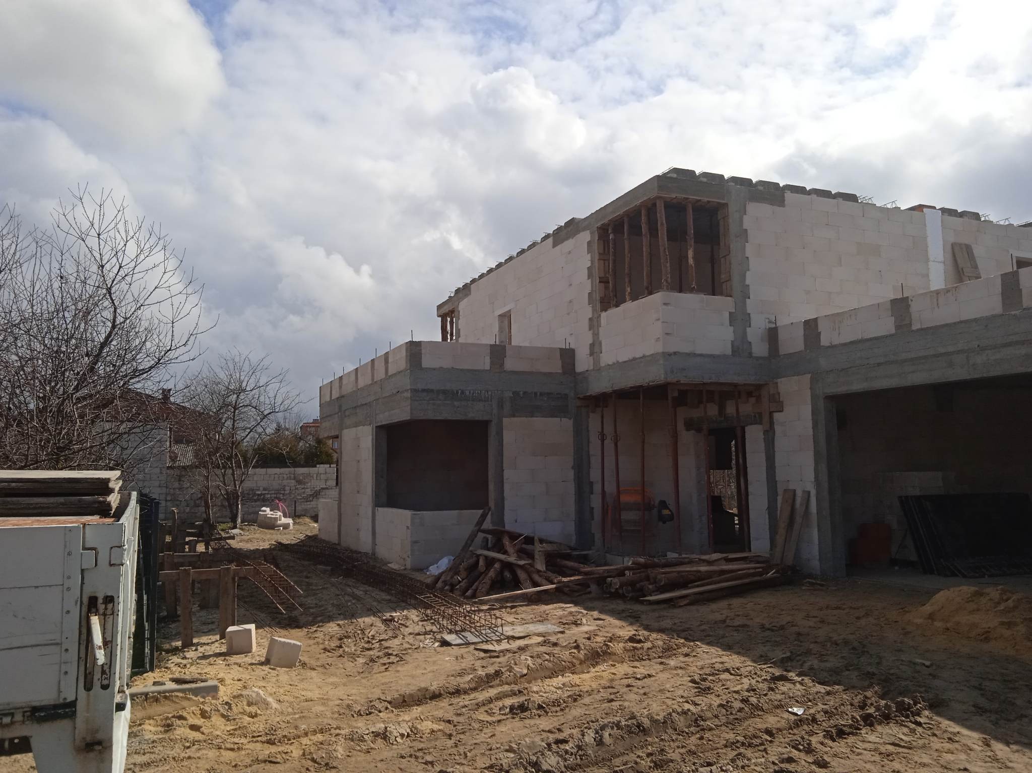 Surowy stan domu z białego bloczka, widoczne zbrojenia i drewniane podpory. Częściowo zarysowany krajobraz z drzewami i pochmurnym niebem w tle. Etap budowy.