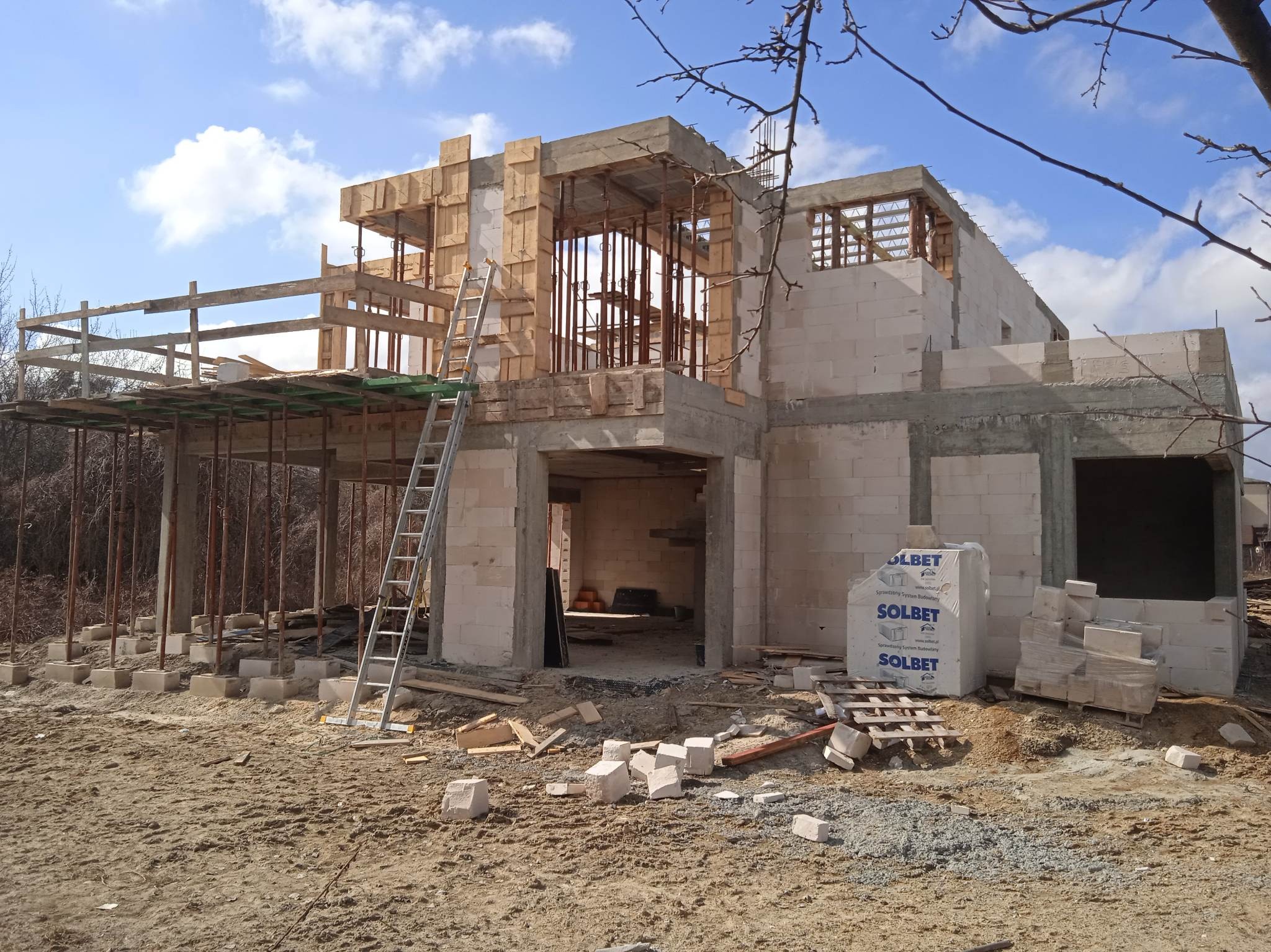 Budowa domu jednorodzinnego w stanie surowym otwartym. Widoczne białe bloczki Solbet, drewniane szalunki i stalowe podpory. Na pierwszym planie drabina i gruz budowlany.