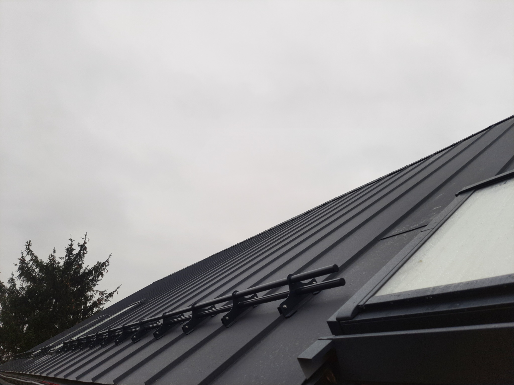 Czarny, blaszany dach z oknem dachowym i systemem odśnieżania, widziany pod kątem. W tle pochmurne niebo i fragment drzewa iglastego.
