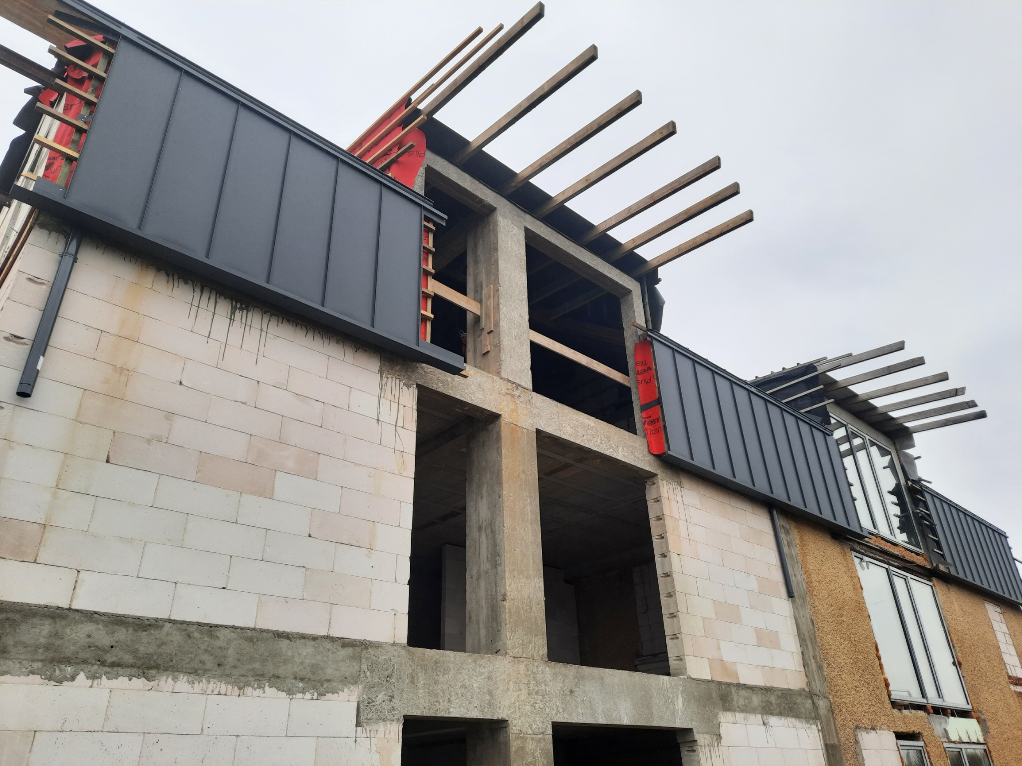 Niedokończony dom z białego pustaka, widoczne betonowe elementy konstrukcyjne, drewniane krokwie i szara blacha na dachu. Ujęcie z dołu, pochmurne niebo w tle.