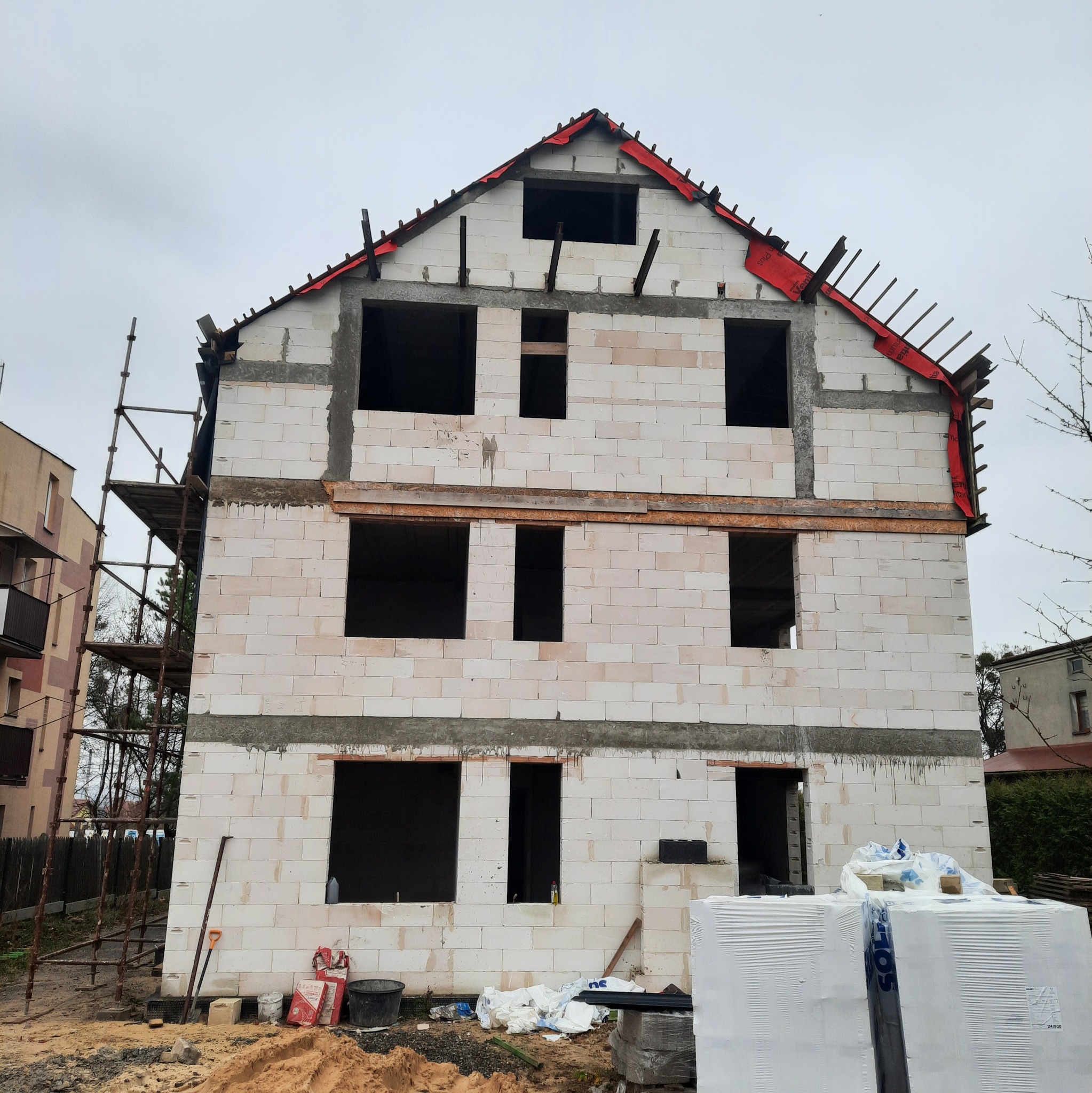 Dwukondygnacyjny dom w trakcie budowy z białych bloczków, widoczne rusztowania i materiały budowlane. Dach zabezpieczony czerwoną folią. Szary dzień.