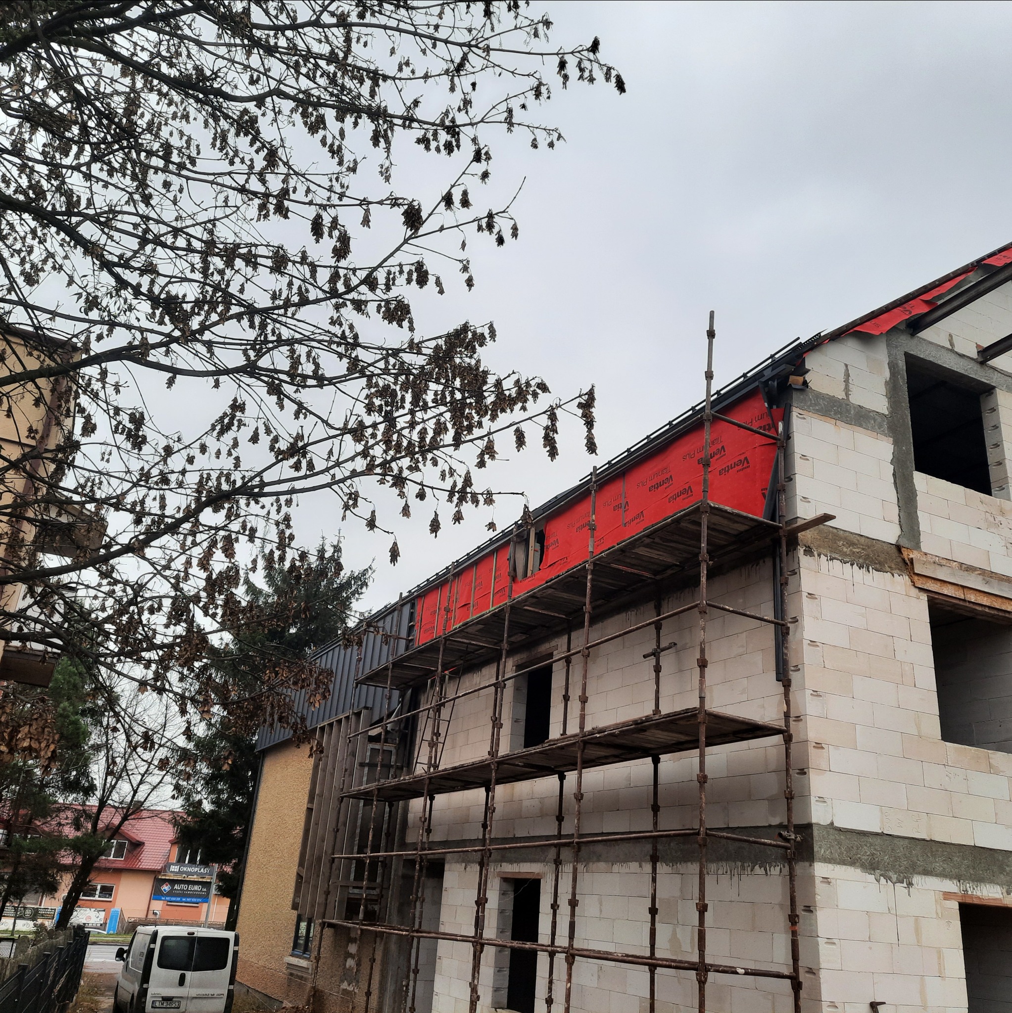 Budowa domu z białych bloczków, widoczne rusztowania i czerwona membrana dachowa. Częściowo ocieplona ściana szczytowa. Drzewo z brązowymi liśćmi na pierwszym planie.