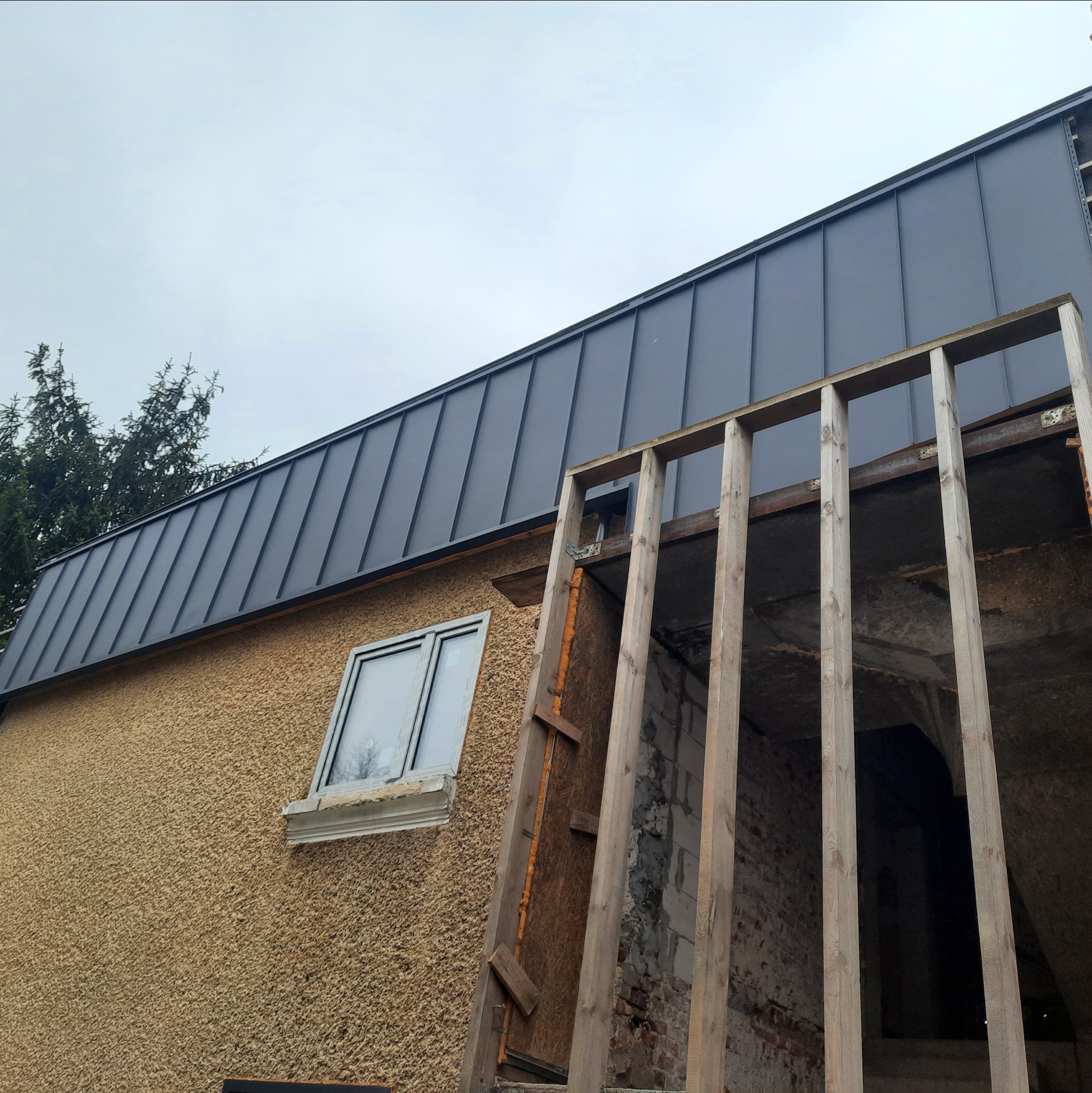 Dom z elewacją o fakturze baranka i nowym, grafitowym dachem z blachy na rąbek stojący. Widoczna drewniana konstrukcja dobudowy.