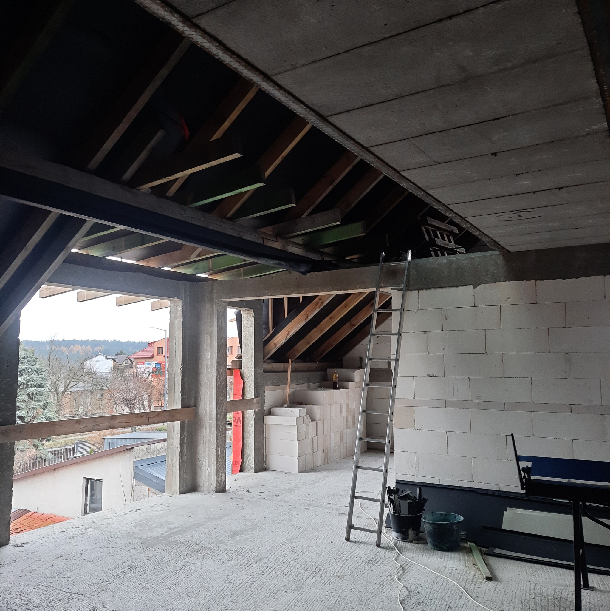 Wnętrze surowego domu w trakcie budowy: ściany z białych bloczków, drewniana konstrukcja dachu, drabina oparta o ścianę. Widok na zewnątrz przez otwory okienne.