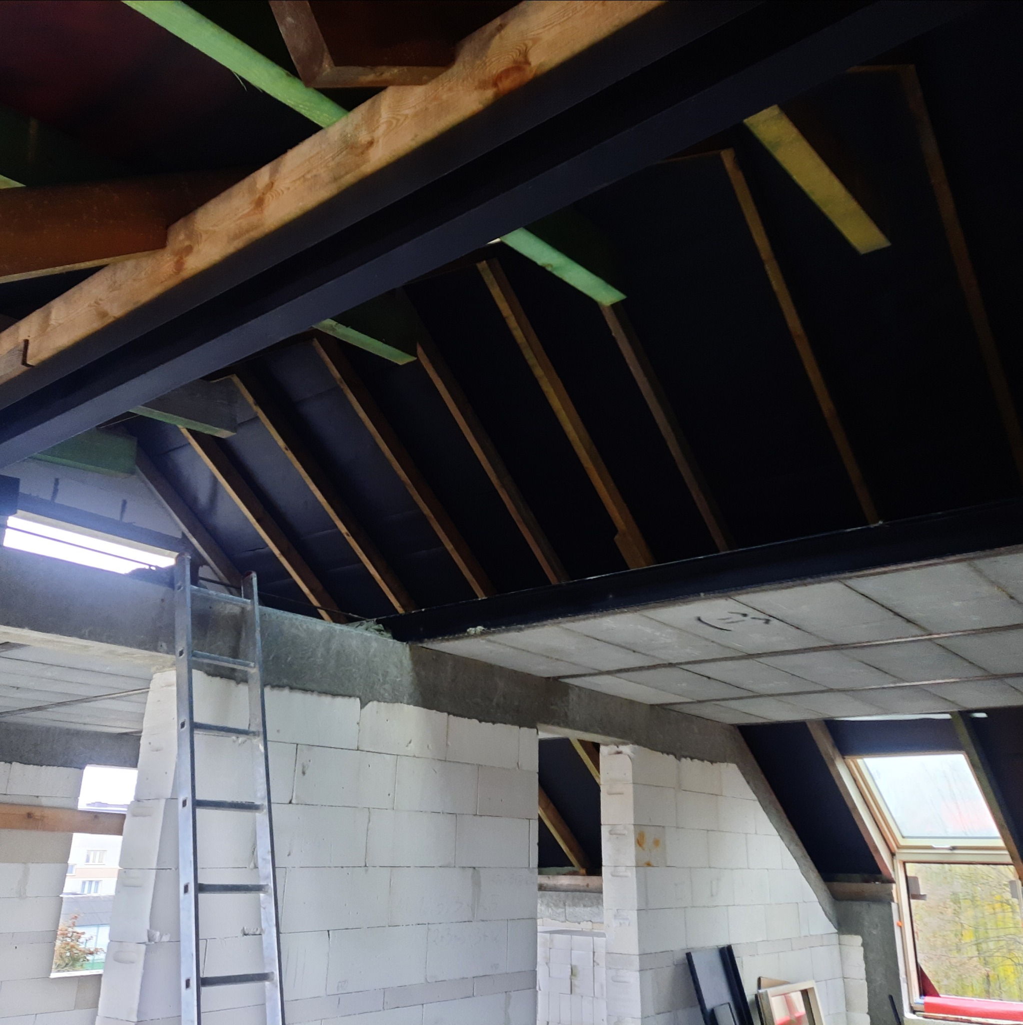 Poddasze w trakcie adaptacji: widoczne krokwie, izolacja, ściany z betonu komórkowego i drabina. Okno dachowe w tle. Etap prac wykończeniowych.