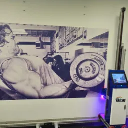 Ściana z czarno-białym muralem kulturysty, obok automat malarski Rayline z niebieskim światłem. Detal wnętrza, potencjalnie po malowaniu.