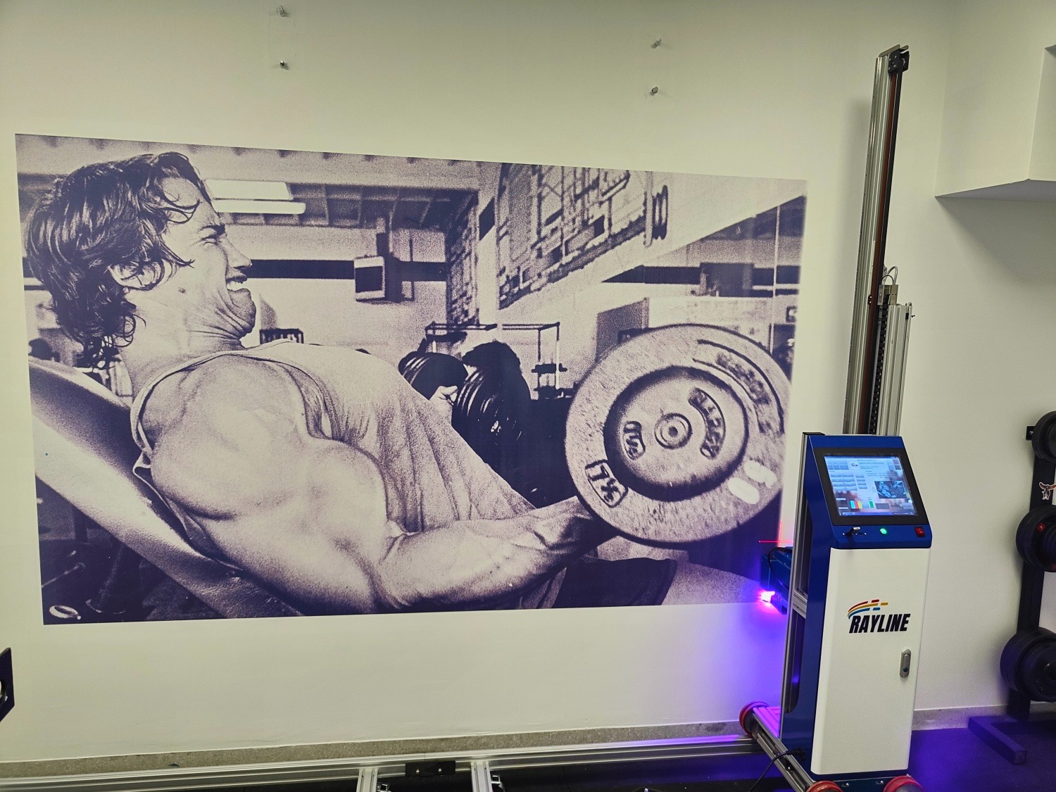 Ściana z czarno-białym muralem kulturysty, obok automat malarski Rayline z niebieskim światłem. Detal wnętrza, potencjalnie po malowaniu.