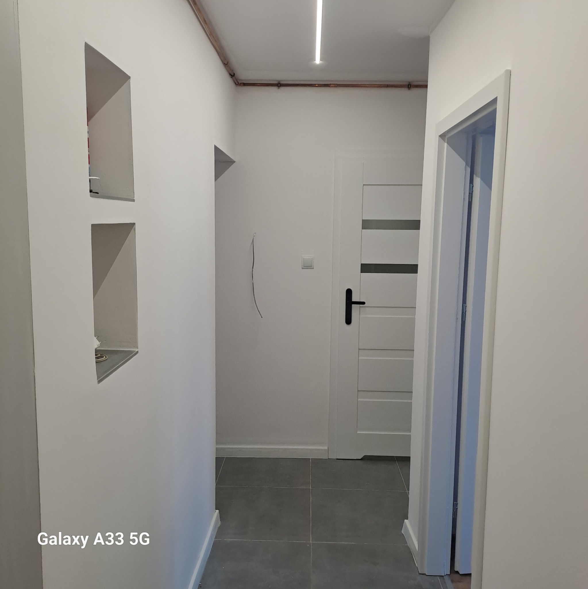 Białe, świeżo pomalowane wnętrze korytarza w Świętochłowicach z wnękami w ścianie, widoczną instalacją pod sufitem i szarą podłogą z płytek.